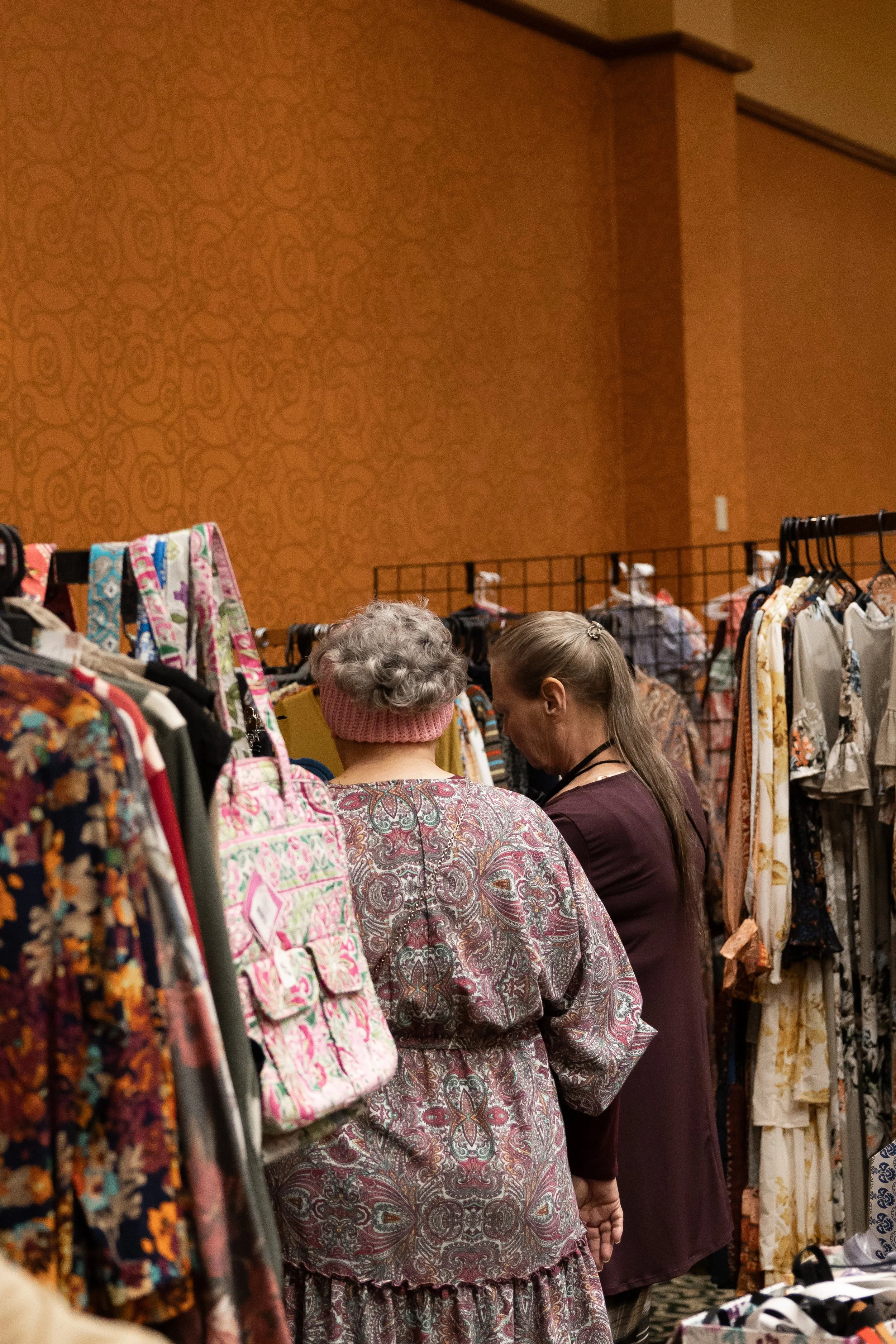 Two women shopping for clothes at a thrift store or boutique, surrounded by racks of clothing and accessories.
