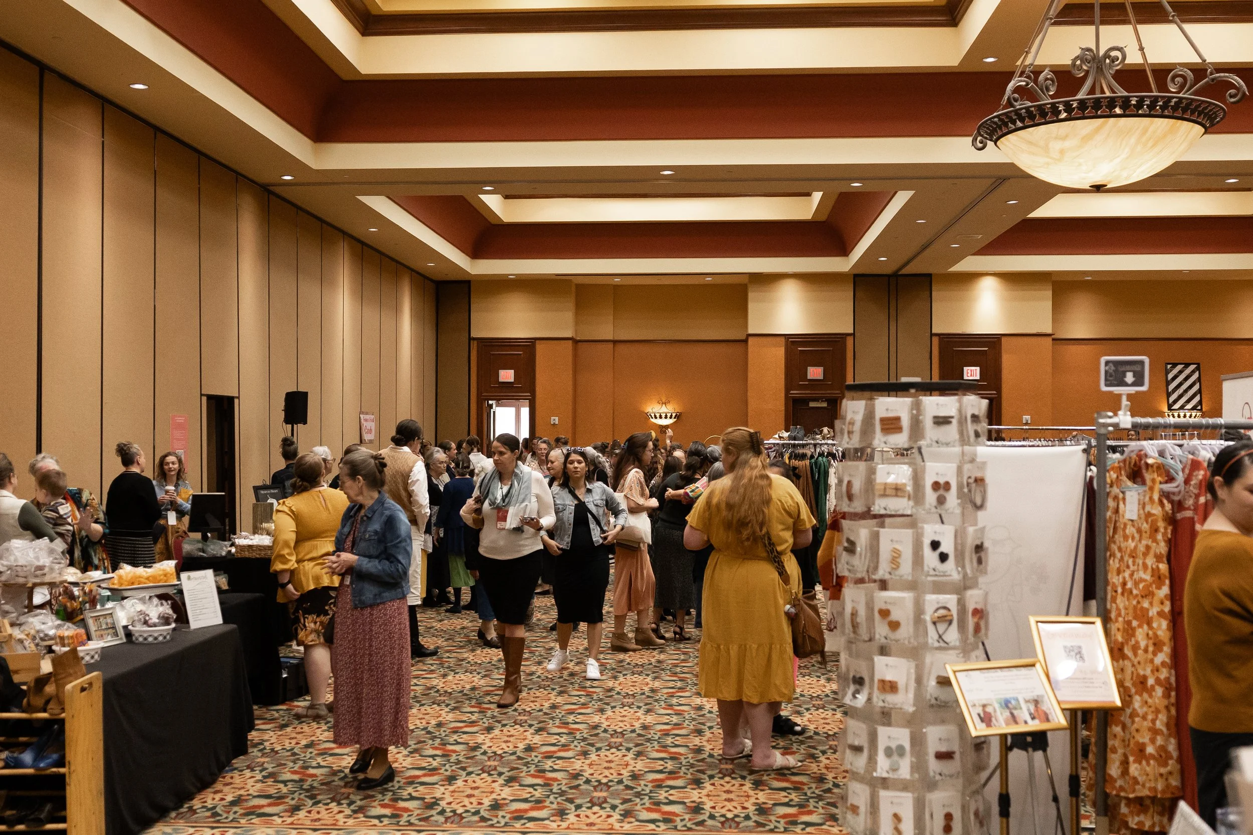 Indoor craft fair with multiple booths and people shopping in a spacious, well-lit room with patterned carpet and high ceiling.