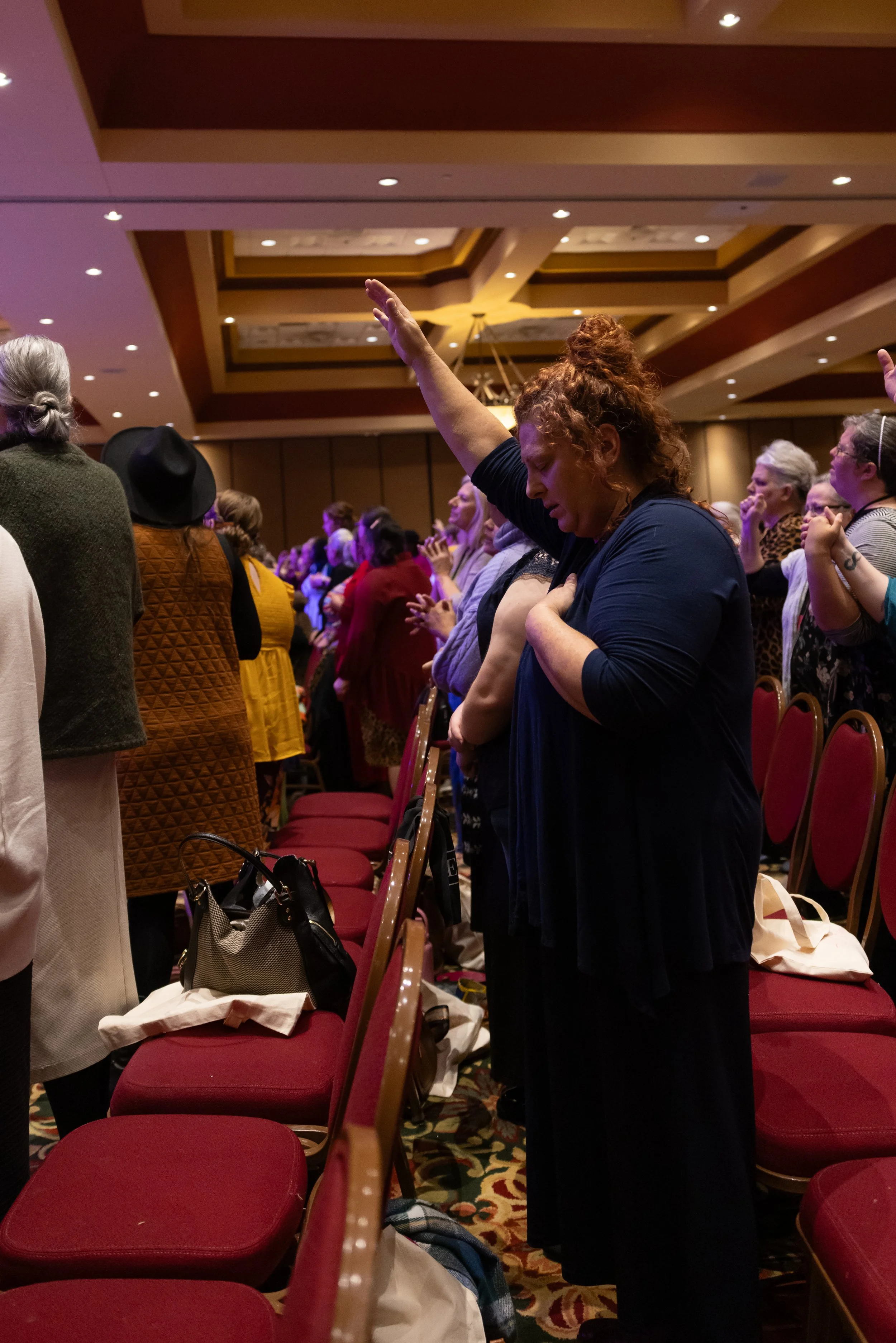 A group of women standing in a prayerful posture with one arm raised inside a conference hall with red chairs and dim lighting.