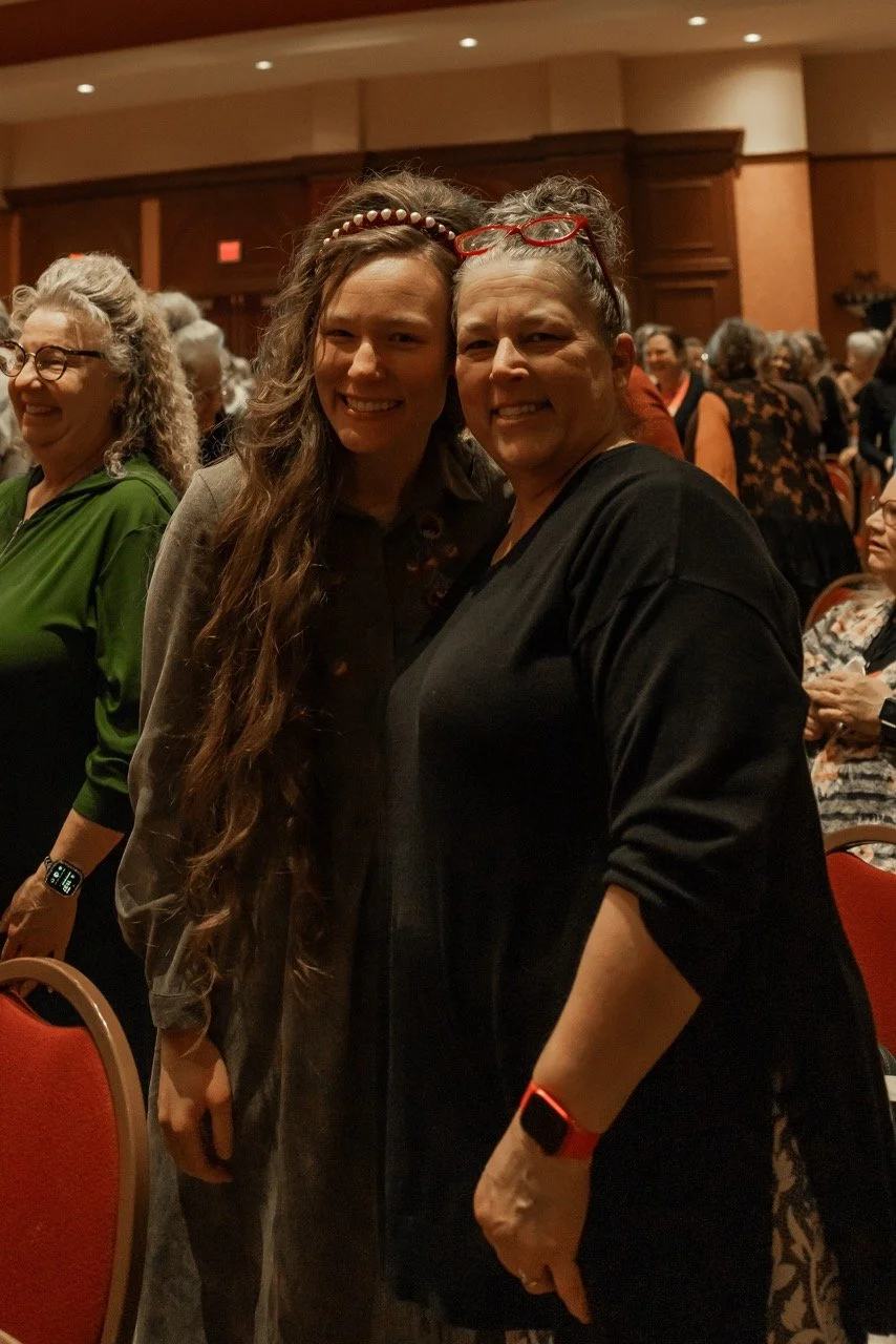 Two women standing close together and smiling at a crowded indoor event with many people in the background.