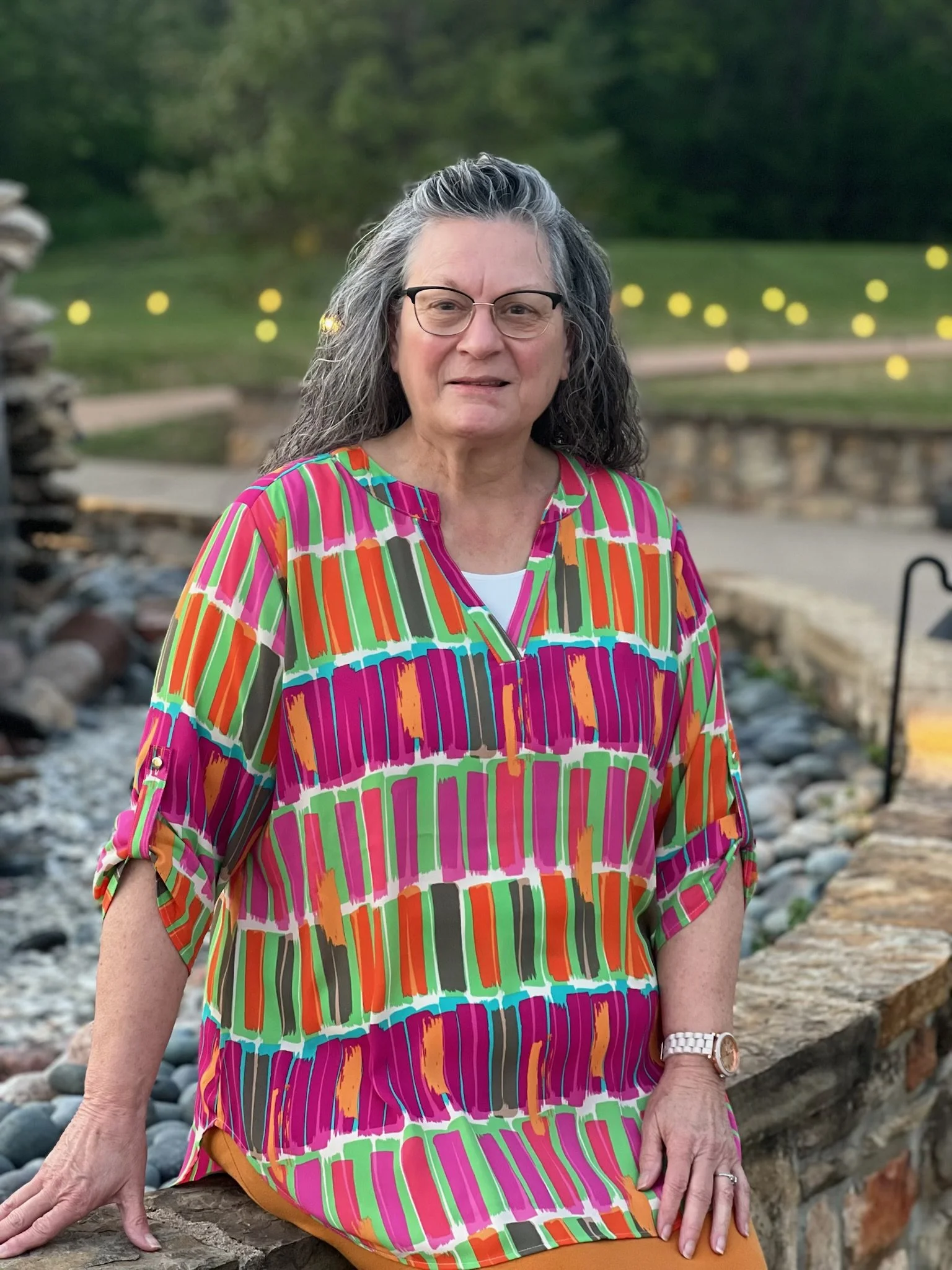 A woman with long gray hair, glasses, and a colorful striped dress sitting outdoors near a stone water feature.