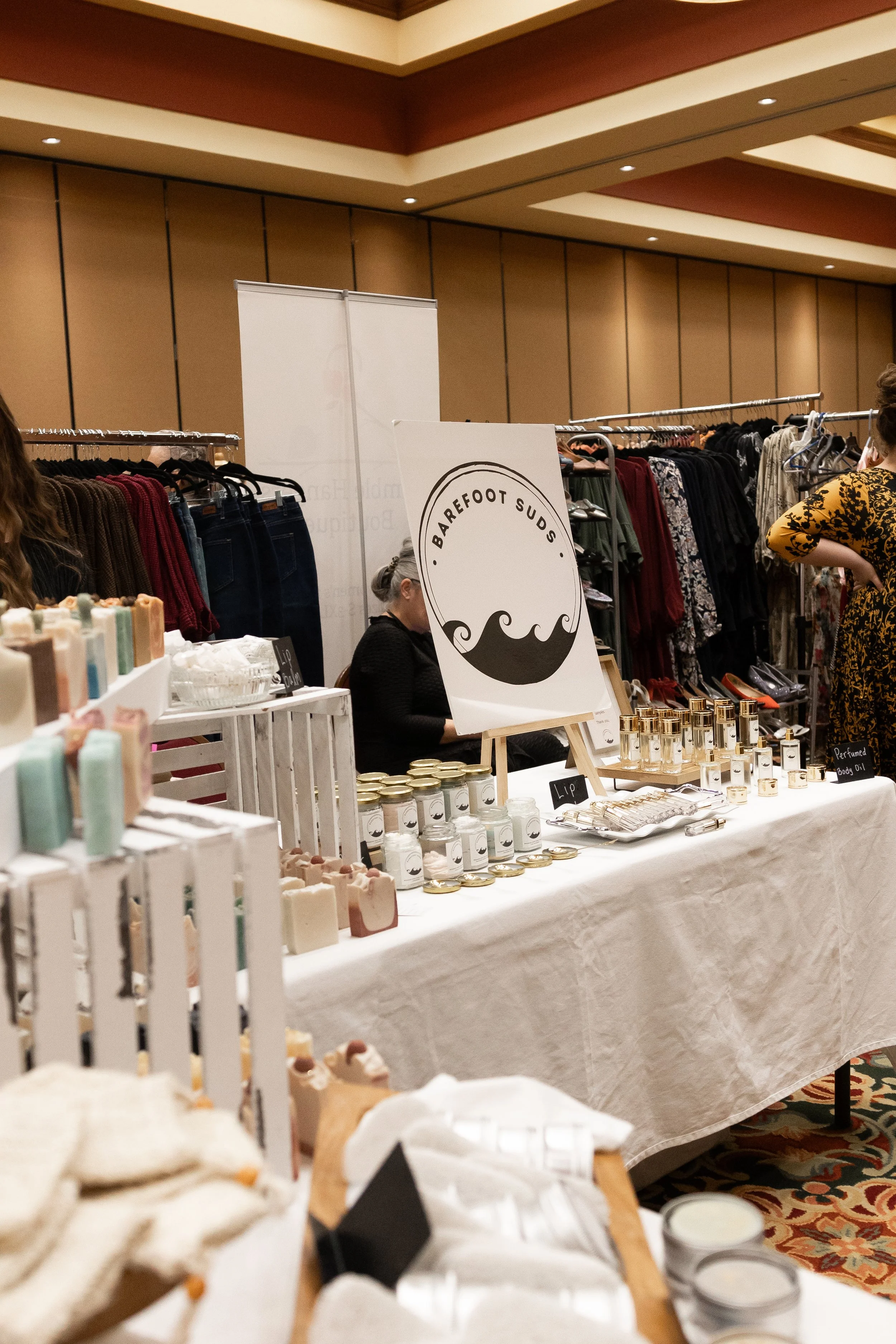 A booth at a market selling natural skincare products with a sign that says 'Barefoot Suds'.