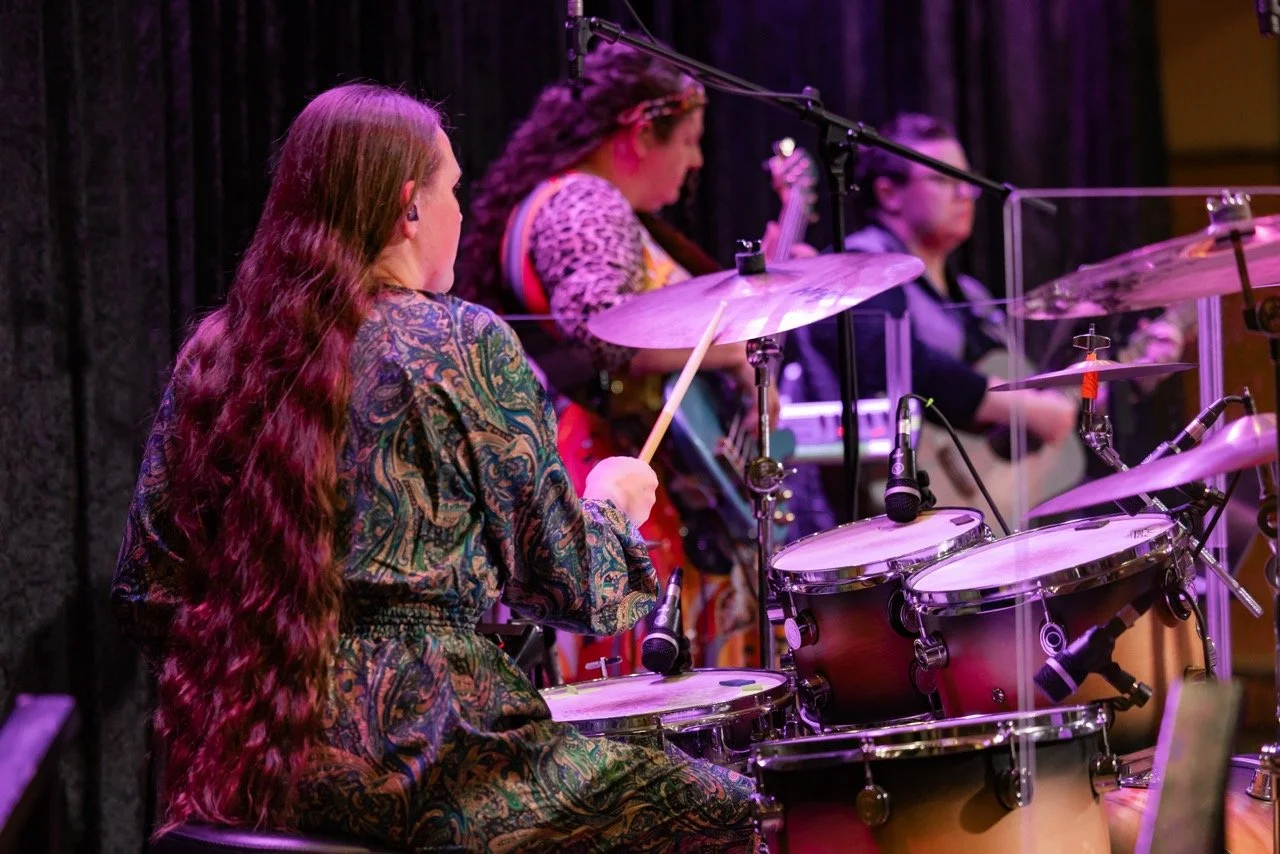 Three women playing musical instruments on stage, with one playing drums in the foreground, another playing bass guitar, and the third holding an acoustic guitar, all dressed in colorful clothing with a black curtain backdrop.