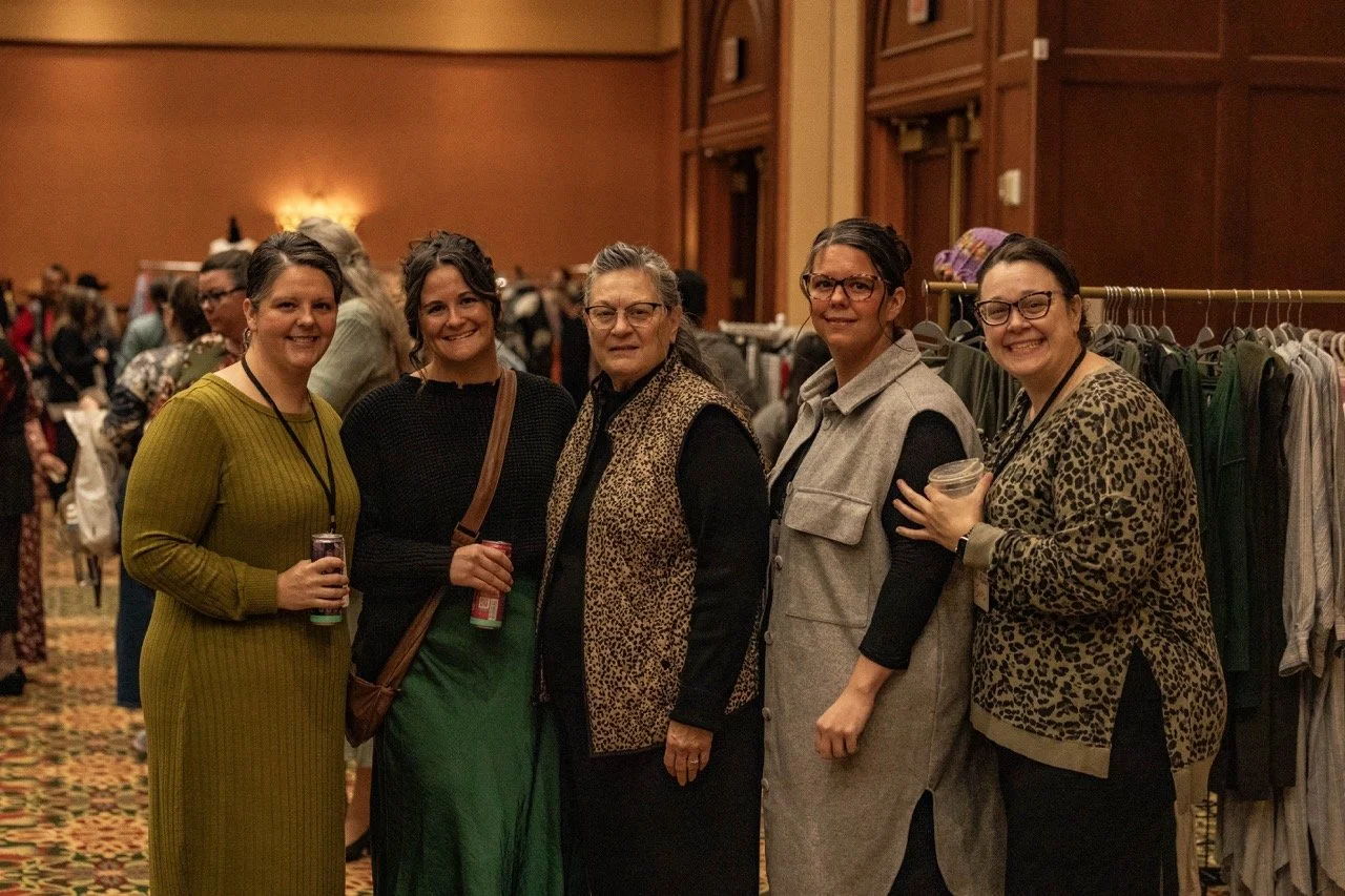 Five women standing together at an indoor event, smiling at the camera, with racks of clothing in the background.