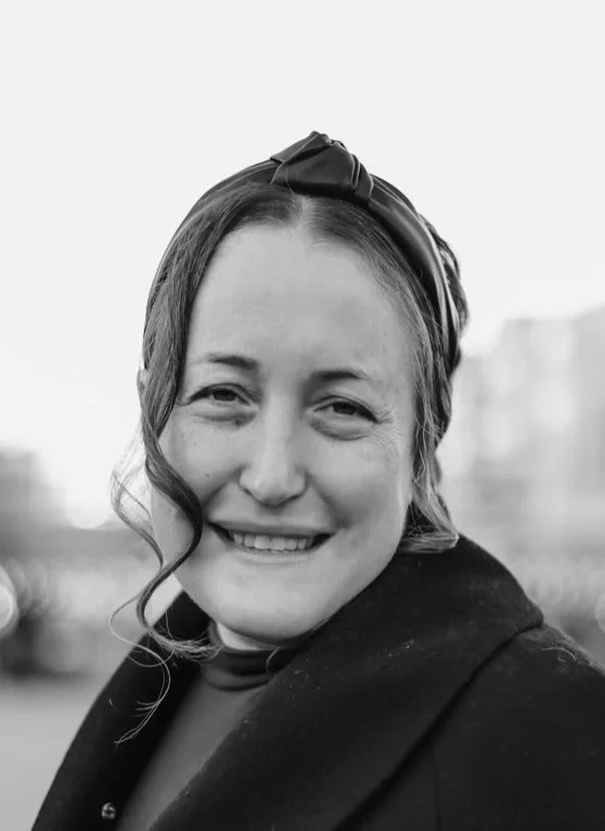 Black and white close-up portrait of a smiling woman wearing a headband and a dark coat, outdoors, with blurred cityscape background.