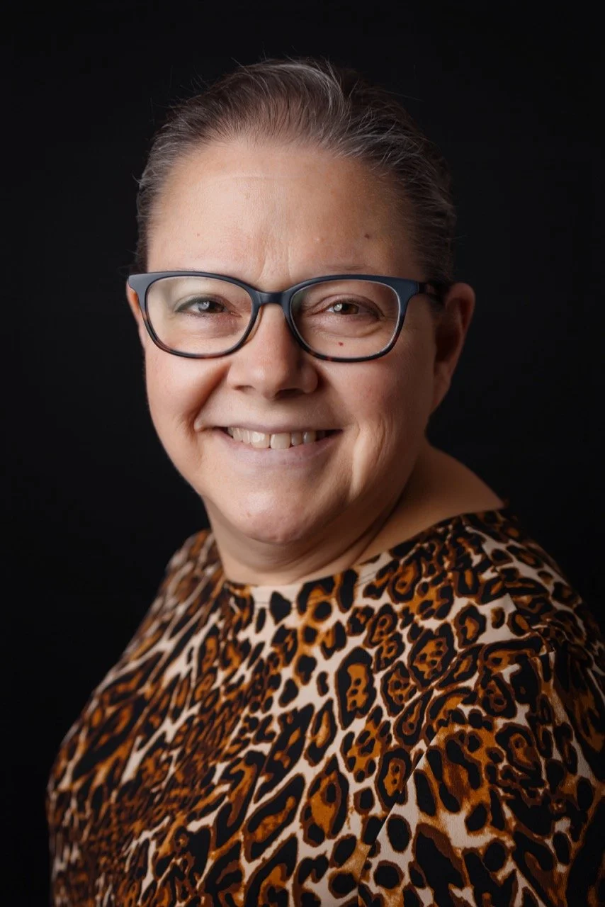 A woman with glasses smiling, wearing a leopard print top, against a black background.