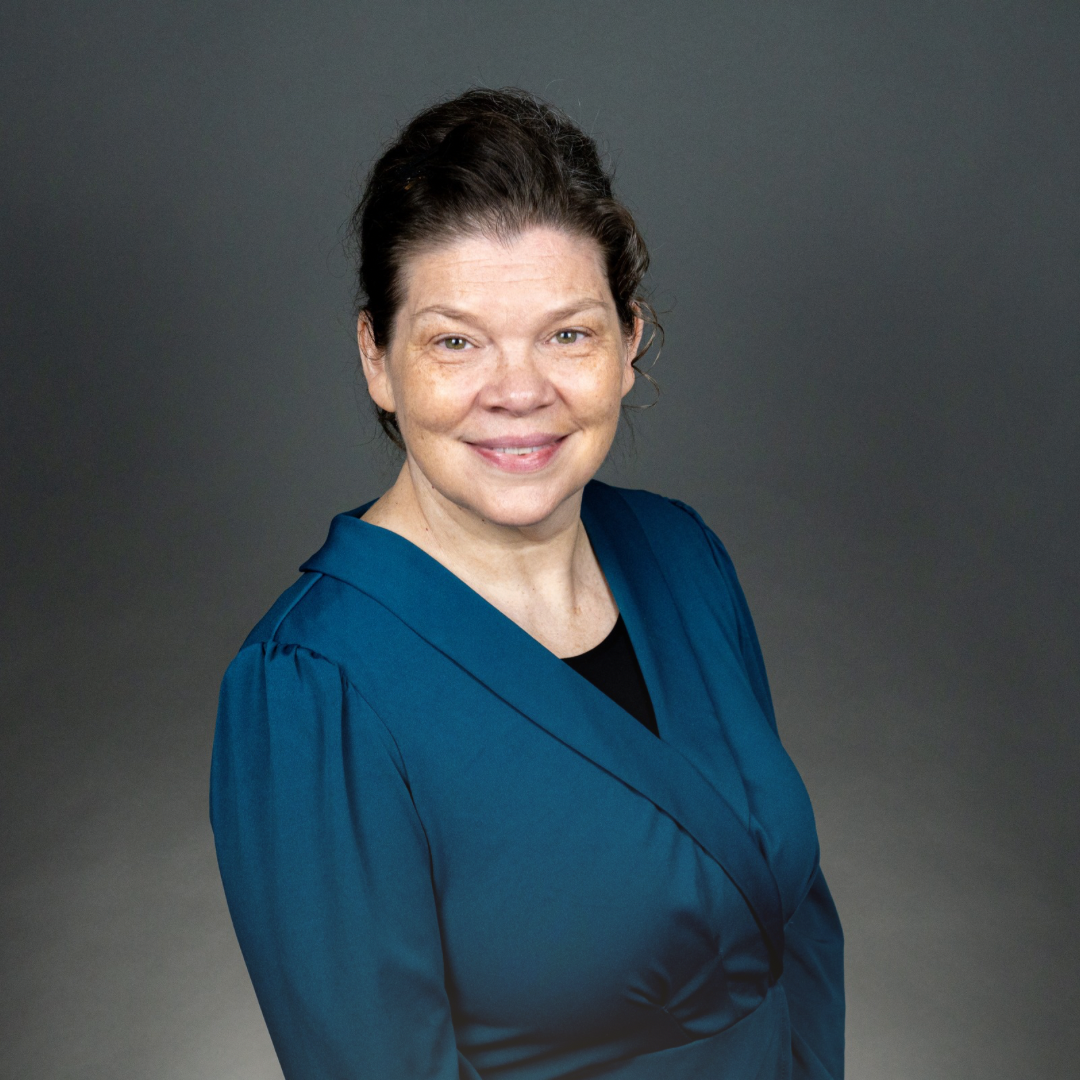 Portrait of a smiling middle-aged woman with dark hair in a ponytail, wearing a blue blazer over a black top, standing against a gray background.