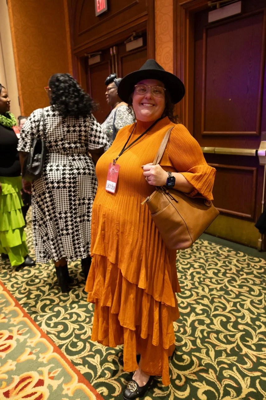 A woman wearing a black hat, glasses, an orange ruffled dress, and black shoes standing on a patterned carpet at a social event, carrying a brown handbag and smiling at the camera.