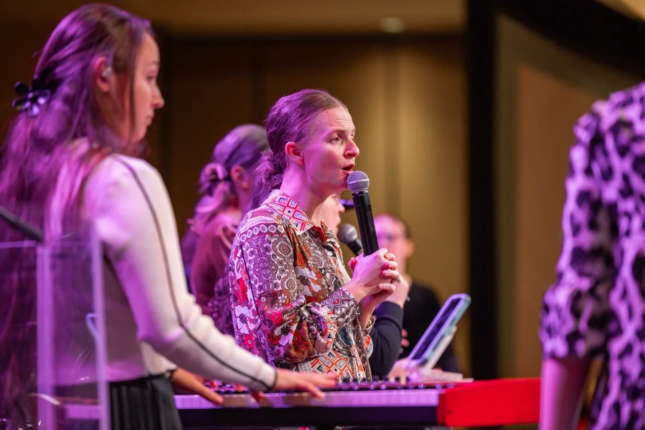 A woman holding a microphone speaking at a conference or event, surrounded by other women, some of whom are looking down at a keyboard or tablet, with purple lighting in the background.