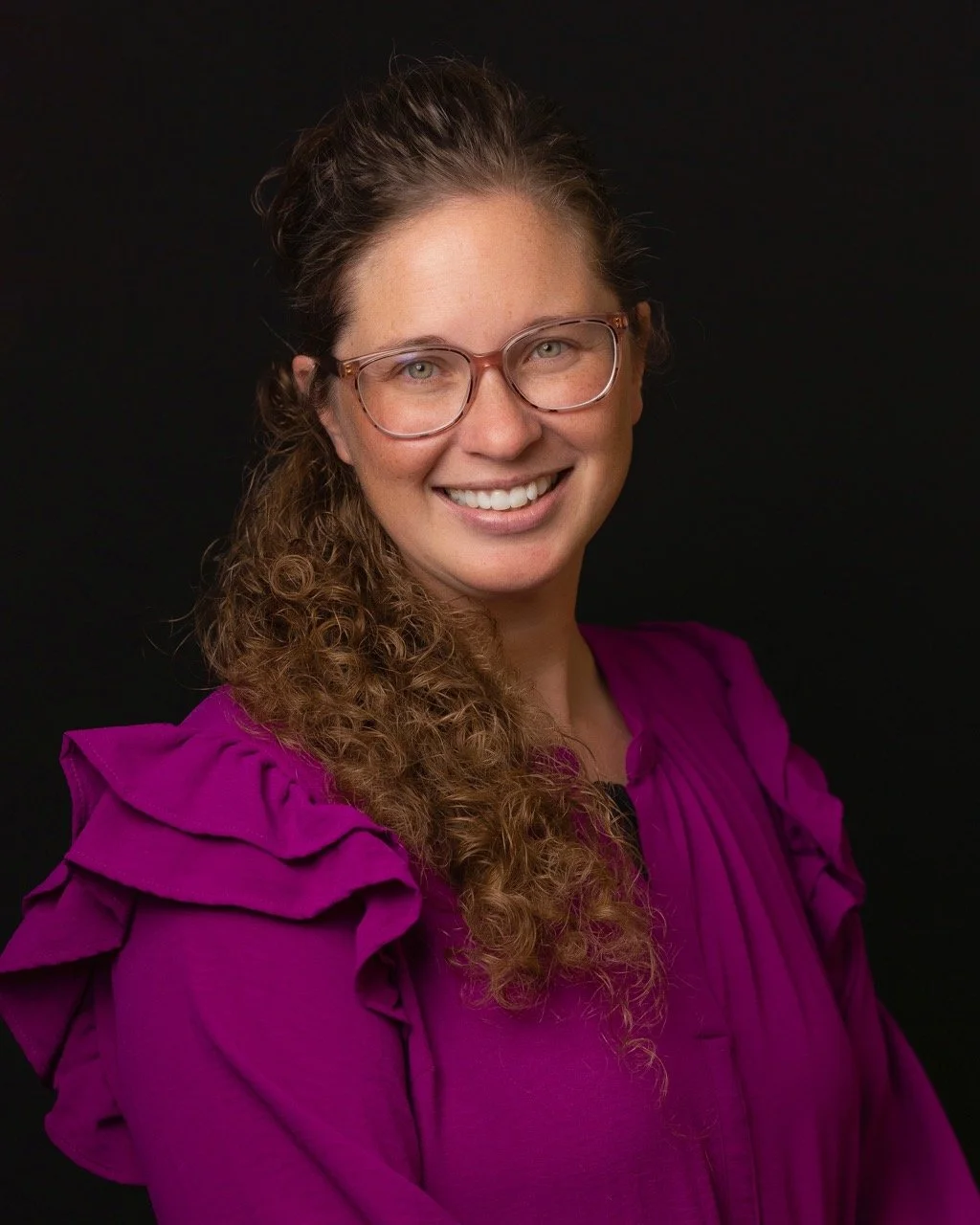 A woman with long curly hair, wearing glasses and a bright purple blouse, smiling against a black background.