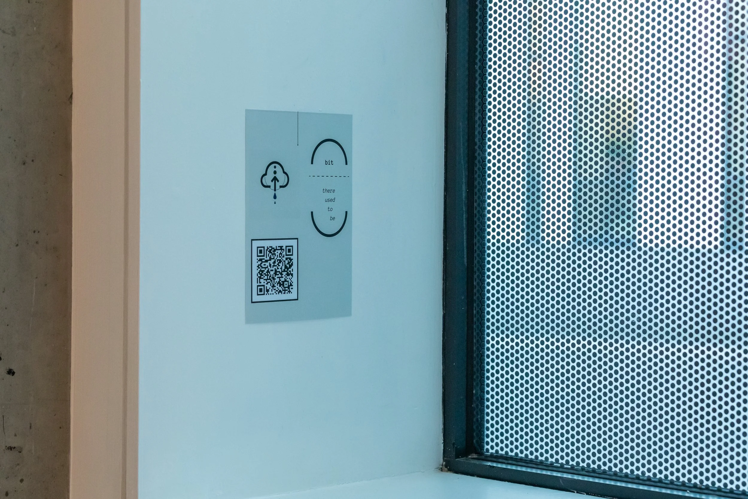 A QR code stuck on a wall by a window. There is a cloud image just above and text that reads: 'bit: there used to be'.