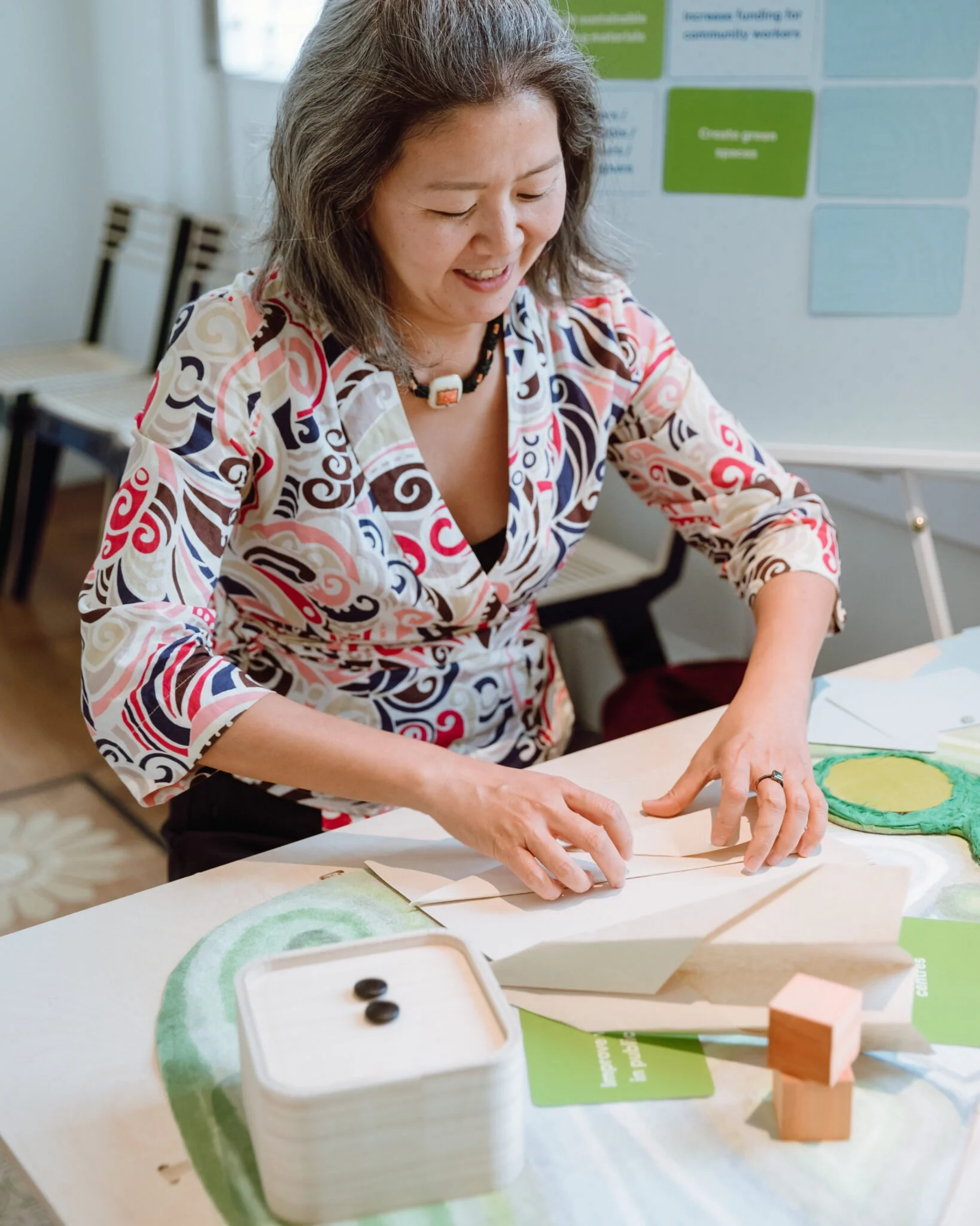 A woman sits folding paper on a table with green fabric and wooden props.