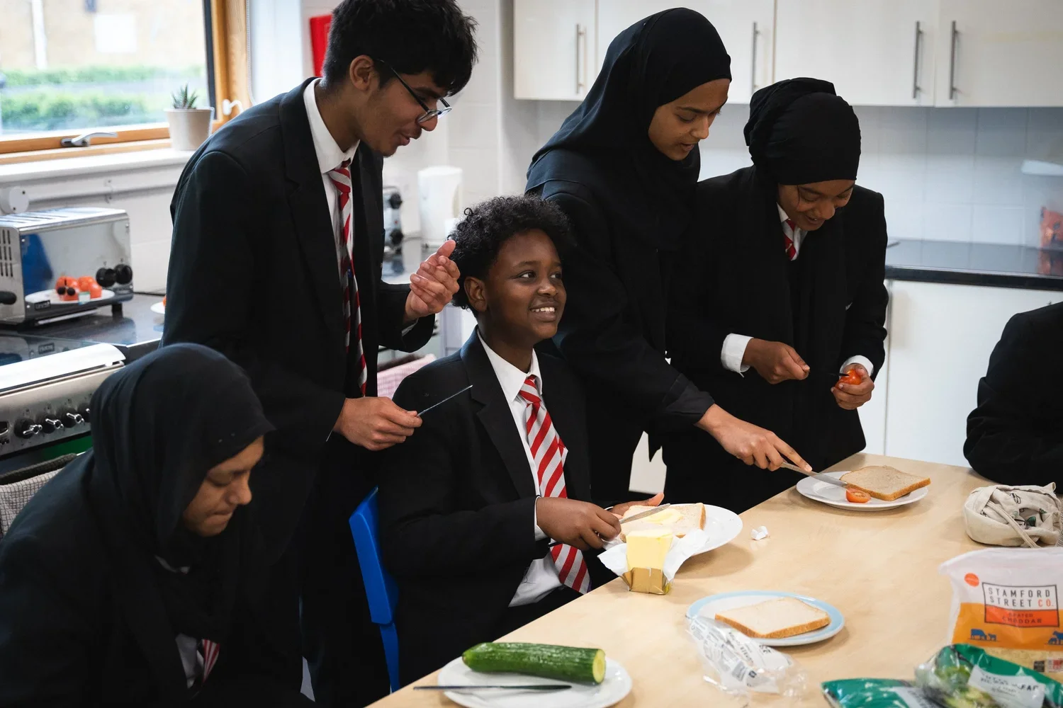 A group of students smile gathered around a table making sandwiches.