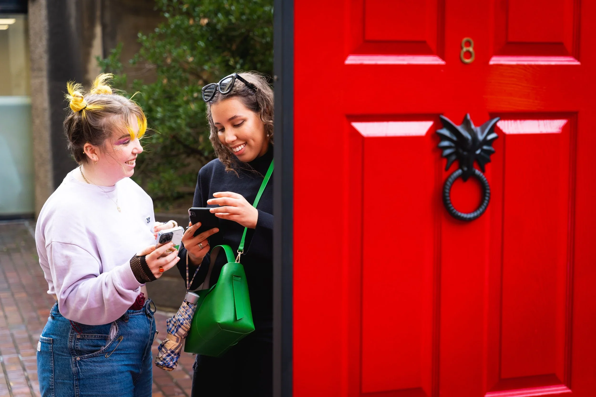 Two people stand smiling, looking at their phones, behind a bright red door.