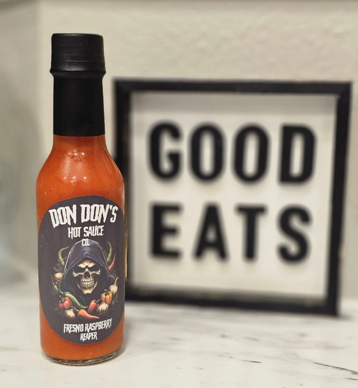 Bottle of Don Don's Hot Sauce Co. Fresno Raspberry Reaper with black cap and label featuring a skeleton in a hoodie surrounded by peppers, in front of a sign that reads 'Good Eats'.