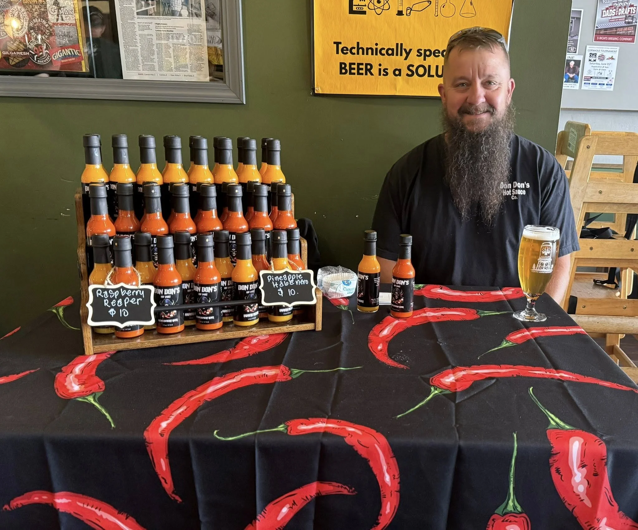 A man with a beard sitting at a table with hot sauce bottles, a glass of beer, and small chalkboard signs displaying flavors like raspberry reaper and pineapple habanero. The table has a chili pepper themed tablecloth, and a yellow sign behind him th