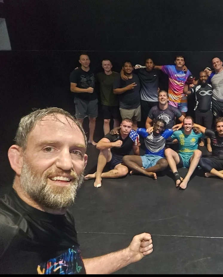 Ryan Long and students at Infinity Martial Arts, some in athletic gear, some sitting on the floor, with one man taking a selfie in the foreground.