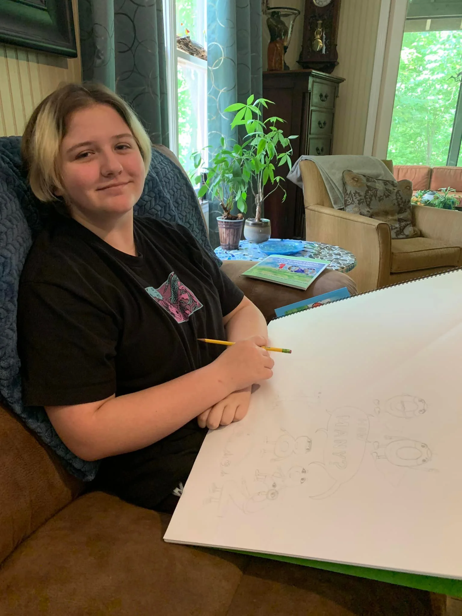 A young girl with short blonde hair sitting on a brown couch in a living room, holding a pencil over a large white sketchbook with a drawing of dogs and the words 'Happy Halloween.' There is a potted plant and a small table with books behind her, and large windows with green trees outside.