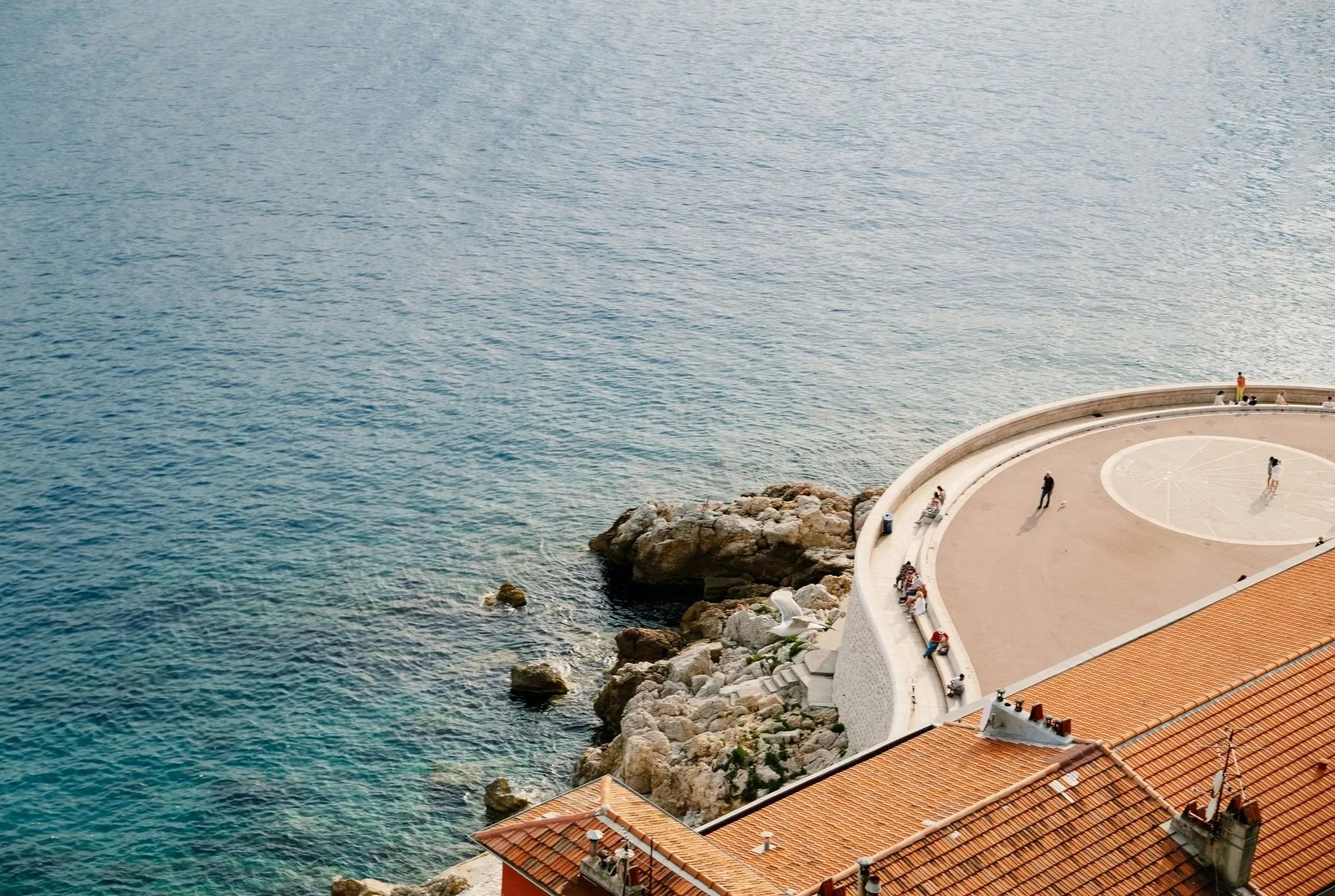 Aerial view of a curved seaside promenade with people sitting, walking, and standing, adjacent to a building with a red-tiled roof, rocky waters and large rocks along the shoreline.