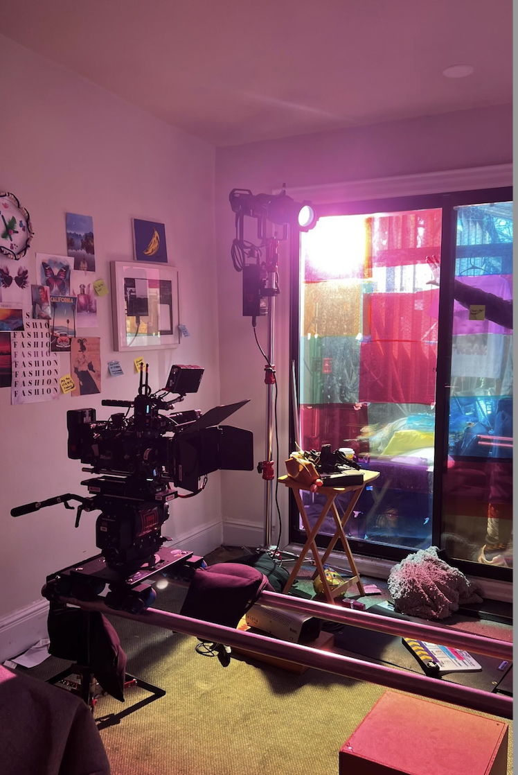 A film or video production setup inside a room with a camera on a stabilizer, studio lights on stands, and a colorful translucent curtain covering a sliding door, with various props, gadgets, and decorations on the floor and walls.