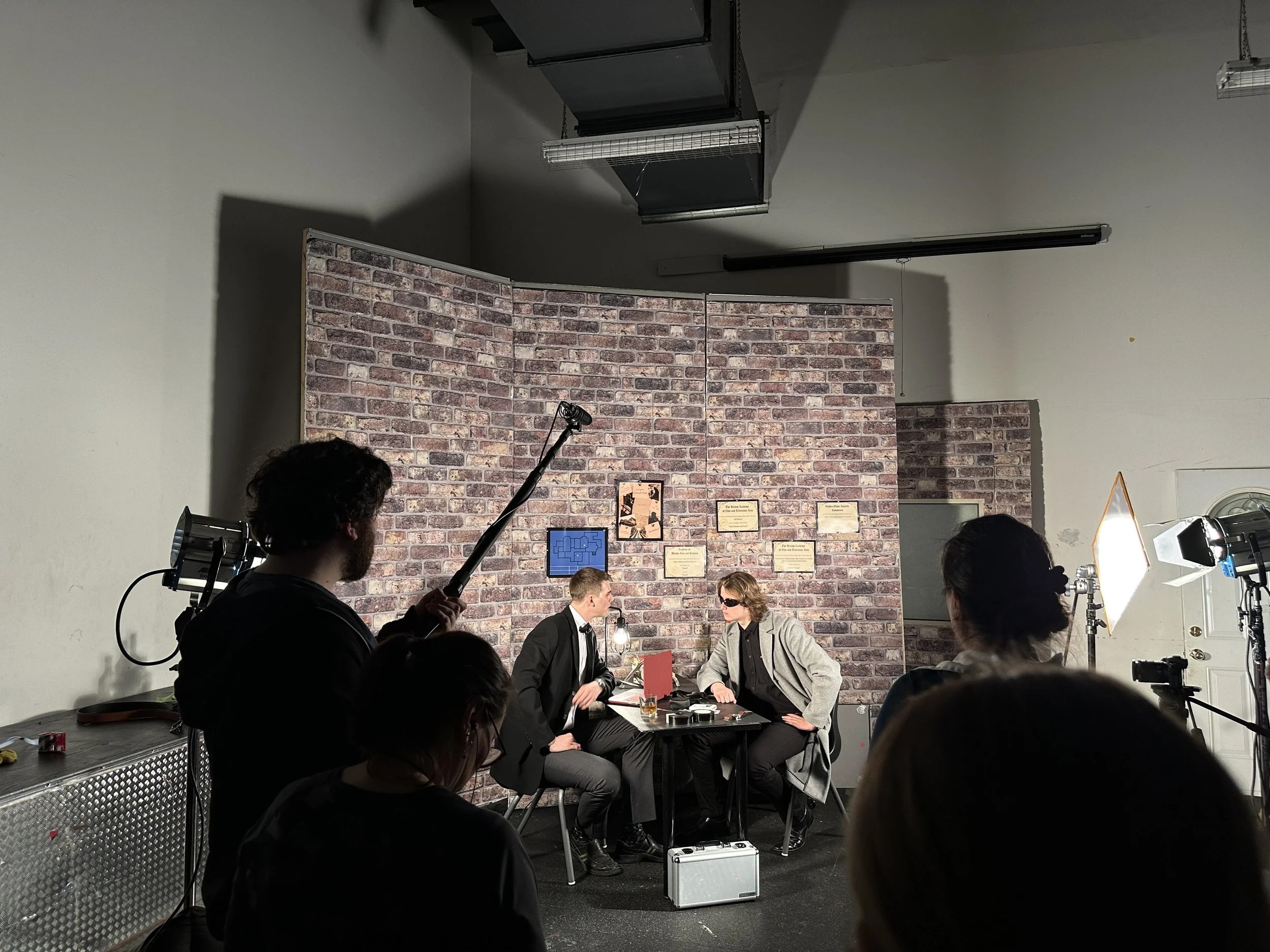 Actors rehearsing a scene in a studio with a brick wall backdrop, cameras, and lighting equipment.