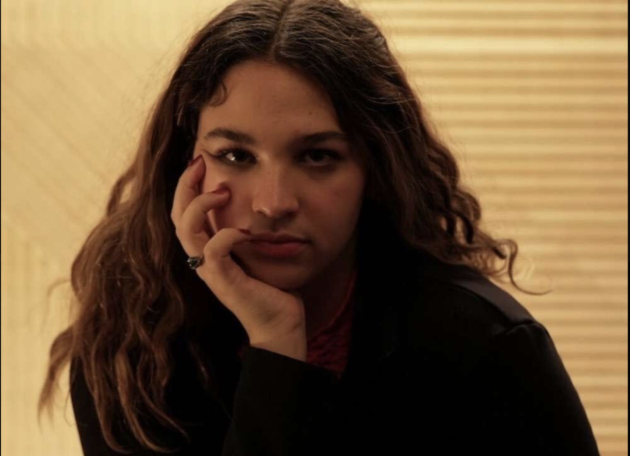 A woman with wavy brown hair resting her face on her hand, looking into the camera with an intense expression, in front of a beige background with wooden blinds.