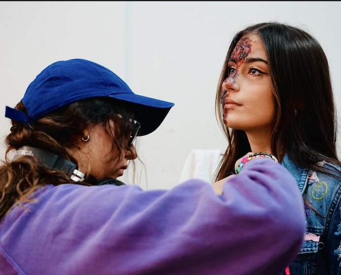 A woman in a blue cap and sunglasses applies makeup or face paint to a young woman's face with a serious expression; the young woman's face has red and black makeup or paint on it.