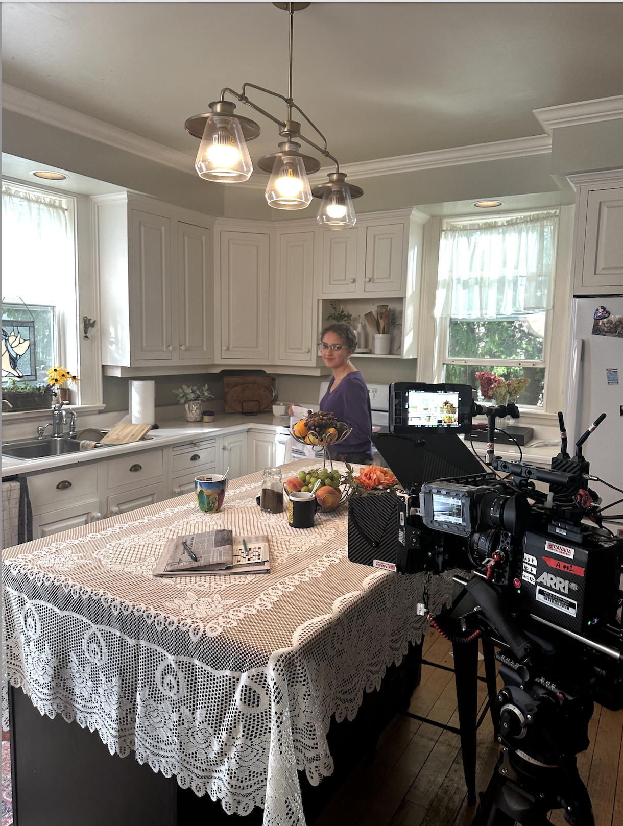 A woman stands in a kitchen with white cabinets, holding a bowl of fruit, with a camera and filming equipment set up on a table covered with a lace tablecloth, in front of her.