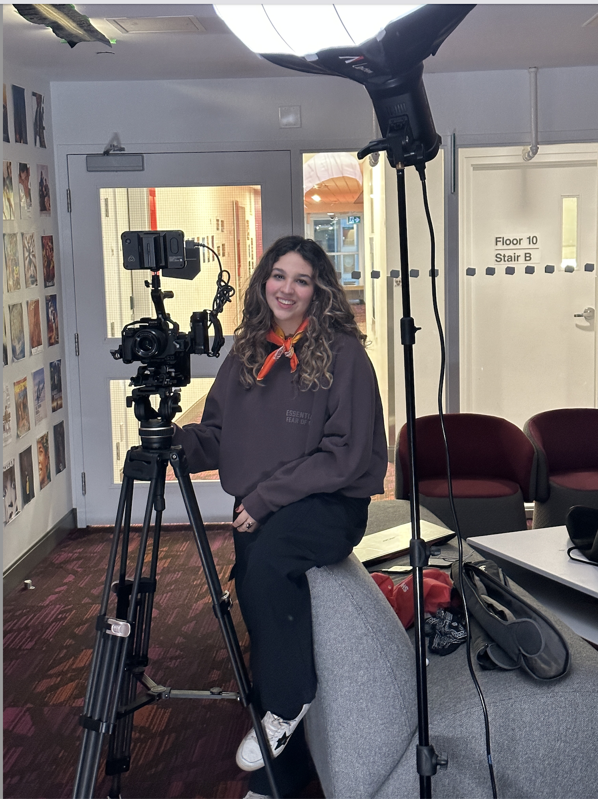 Young woman with curly hair and a red bandana around her neck, sitting beside a camera on a tripod, smiling in a casual indoor setting with chairs, a gray couch, and pictures on the wall.