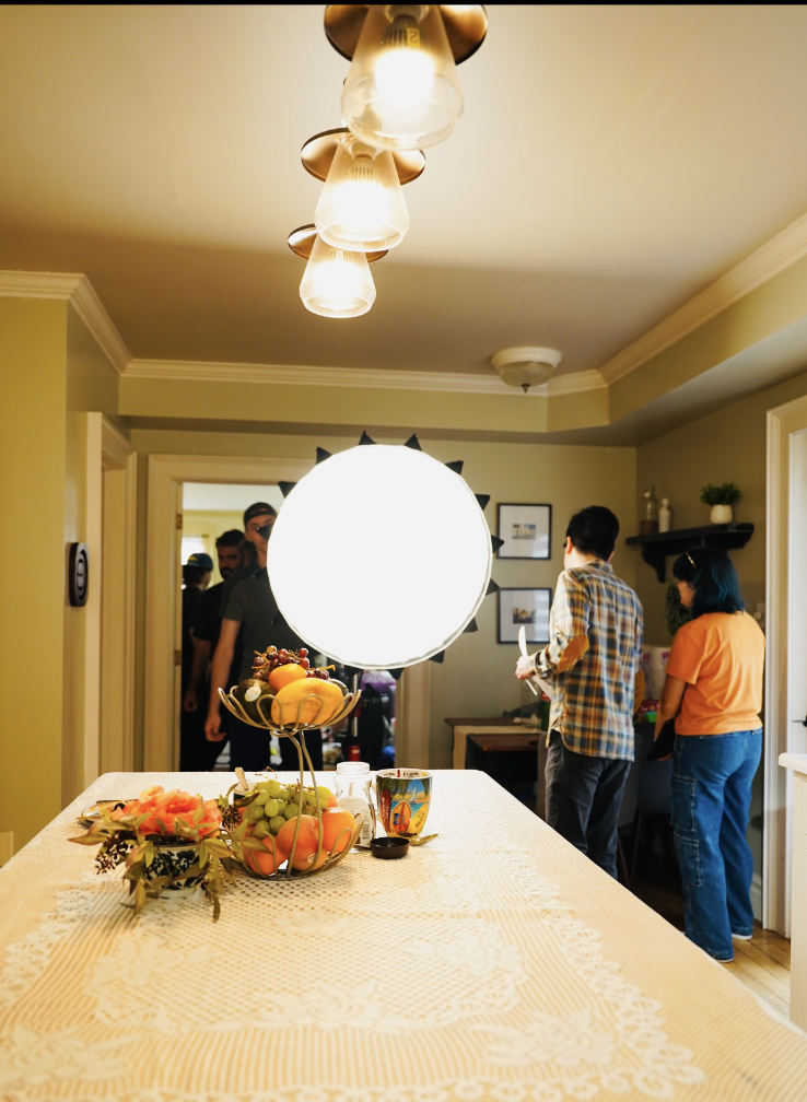Interior of a dining room with a table decorated with a fruit centerpiece and a large bright light in the middle. In the background, several people are gathered near a doorway, some facing away, some partially visible.