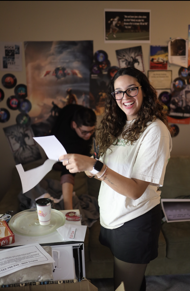 A smiling young woman with glasses and curly hair holding papers, standing in a room decorated with posters and memorabilia on the wall, with a person in the background organizing items.