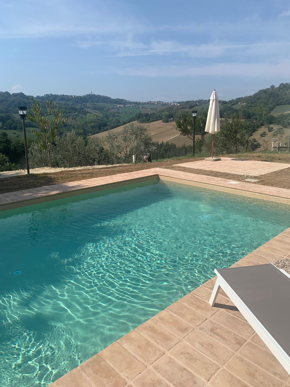 Swimming pool with clear water on a tiled deck, overlooking rolling green hills and a partly cloudy sky, with an open white umbrella and a lounge chair nearby.