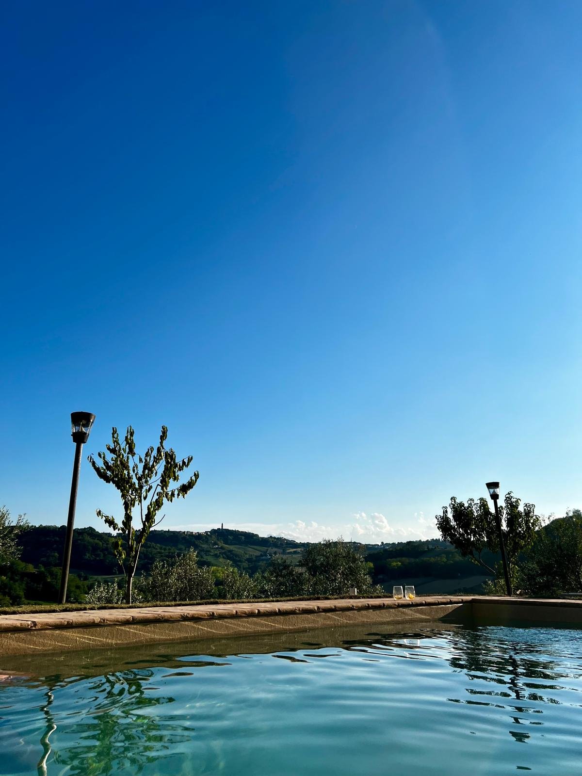 View of a swimming pool with a scenic landscape in the background, including trees, rolling hills, and a clear blue sky.