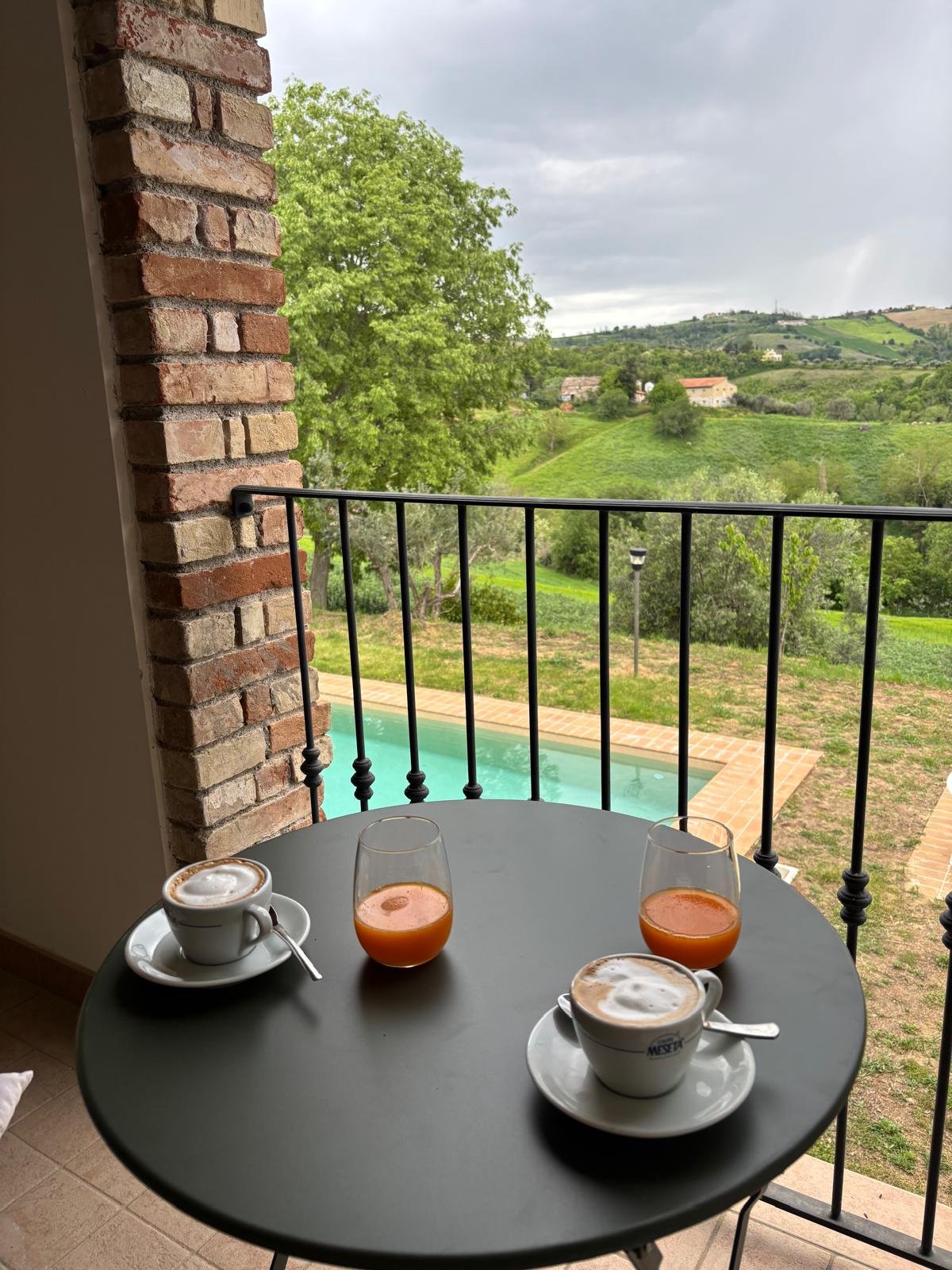 Coffee cups and glasses of orange juice on a balcony table overlooking a landscape with trees, hills, and a swimming pool.