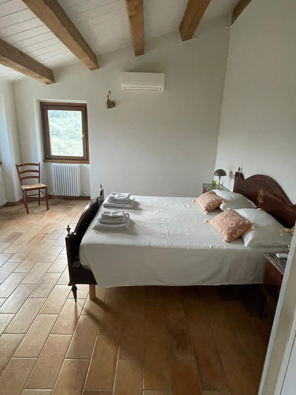 A bedroom with a double bed with white sheets, two pink pillows with a geometric pattern, and folded towels. There's a wooden chair near a window with a radiator underneath it, a nightstand with a lamp, and a window showing outside greenery. The ceil