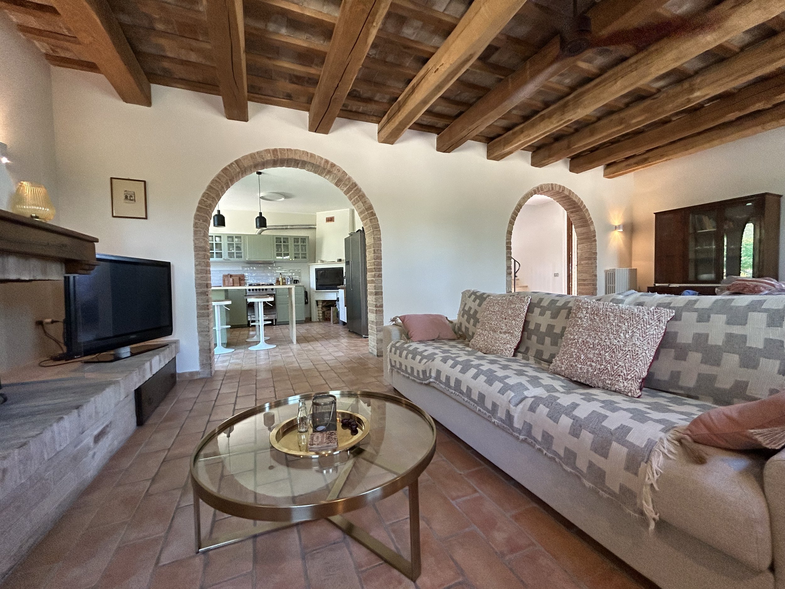 Living room with a brick fireplace, a patterned sofa with multiple pillows, a gold coffee table with drinking glasses, and a view into a kitchen through two arched brick doorways. The ceiling has exposed wooden beams.