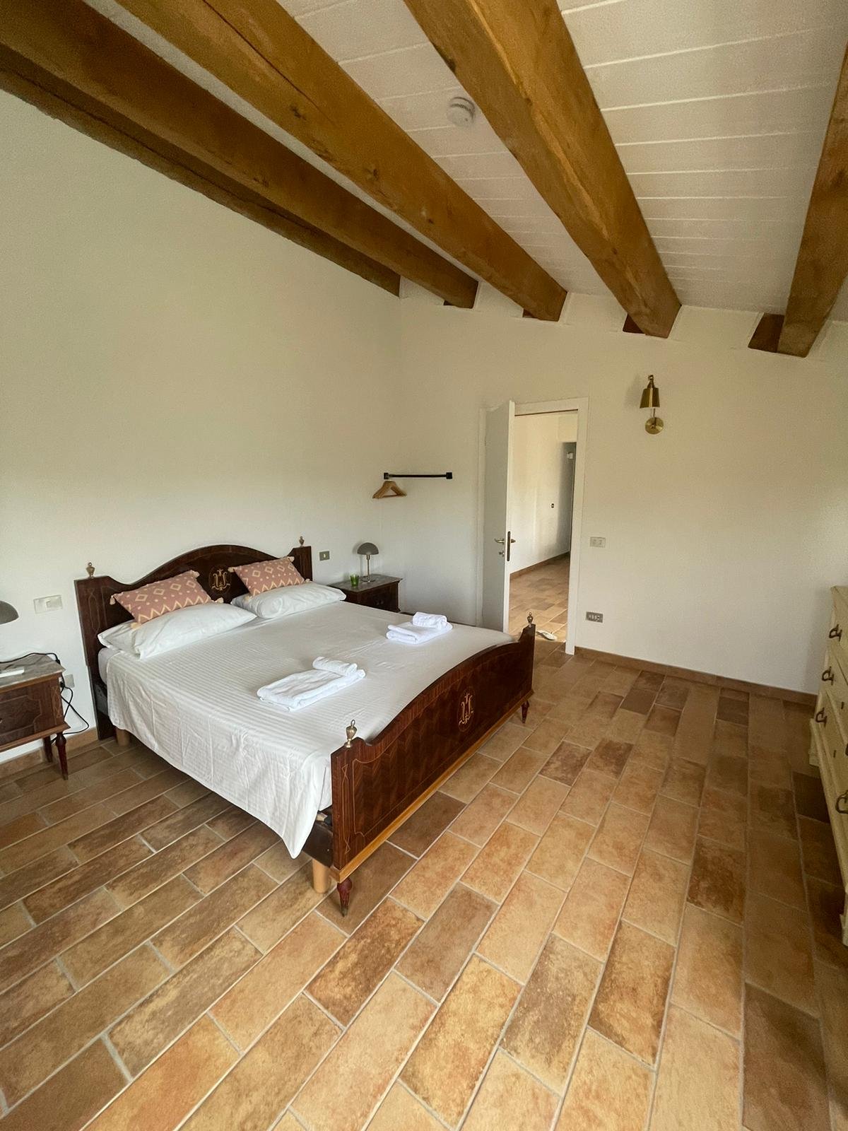 A bedroom with a wooden bed, two bedside tables, and a beige dresser on the right. The room has white walls, wooden beams on the ceiling, and a tiled floor. Towels are folded on the bed, and there is a doorway leading to a hallway.