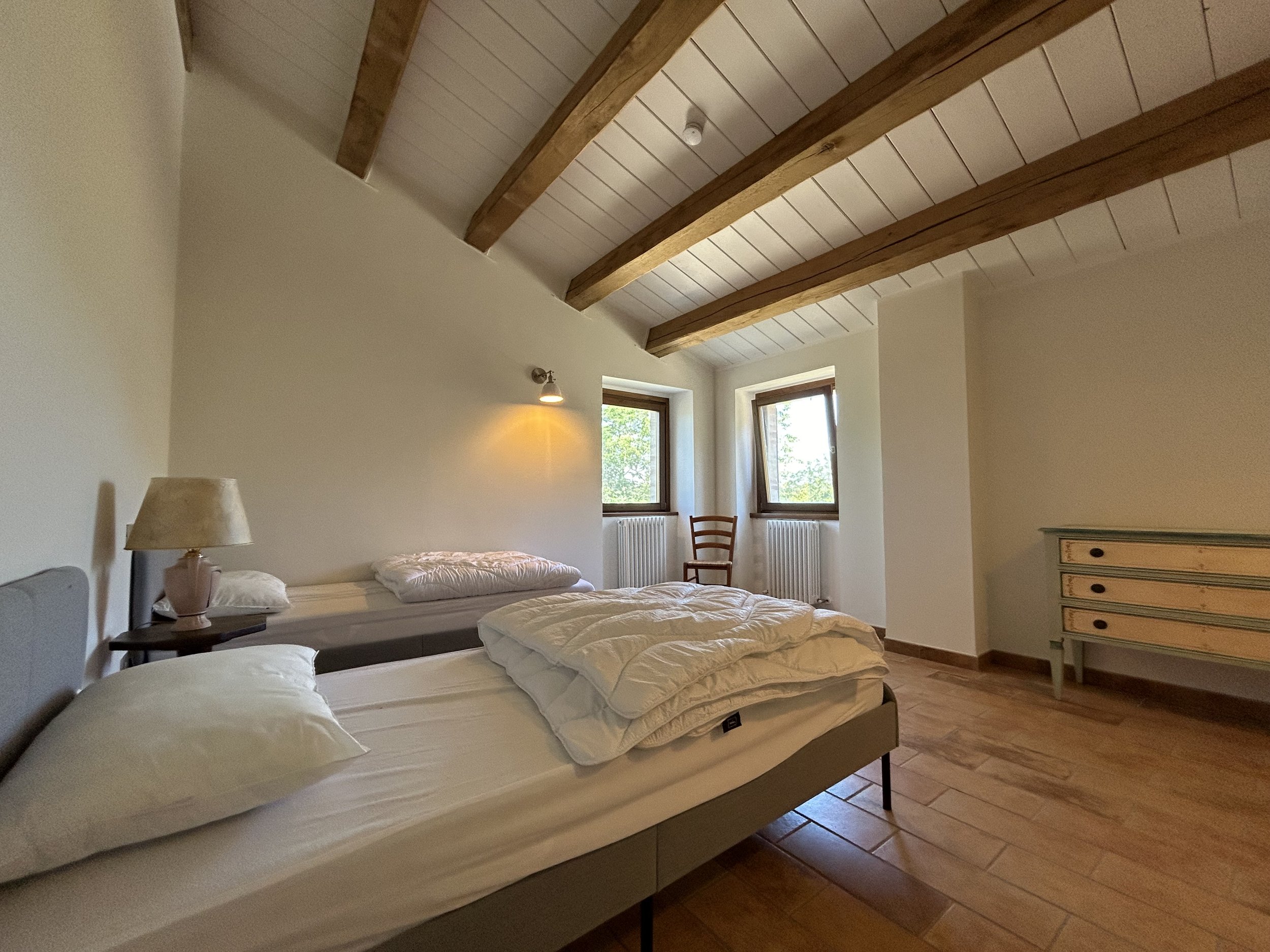 Bedroom with two beds, one with a pillow and blanket, wooden ceiling beams, two windows with sunlight, a small chair by the window, a dresser, and a wall-mounted reading light.