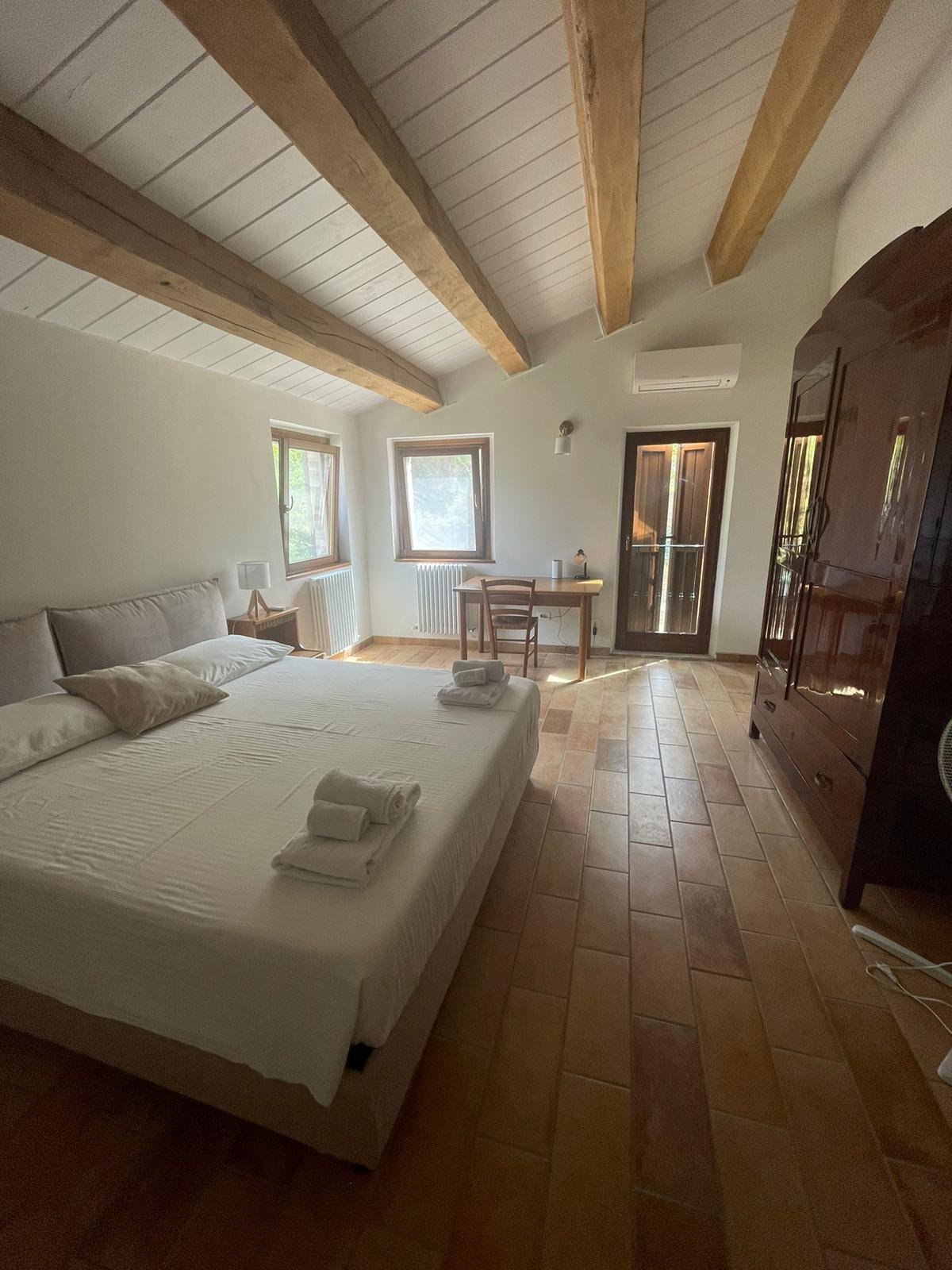 A bedroom with a vaulted ceiling featuring wooden beams, a large bed with white linens and towels, a small desk and chair near the window, a dark wooden wardrobe, and terracotta tile flooring.