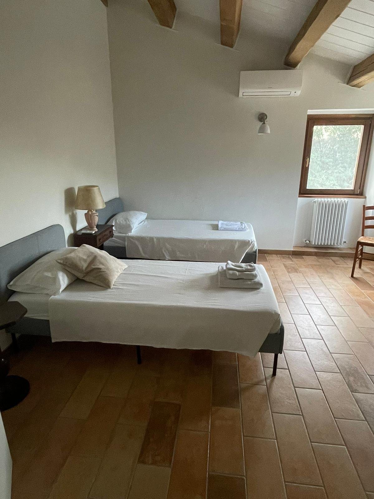 A bedroom with two single beds, one larger and one smaller, with white sheets and pillows. There is a nightstand with a lamp beside the larger bed. A window lets in natural light, and there is a wooden chair and radiator near the window. The ceiling