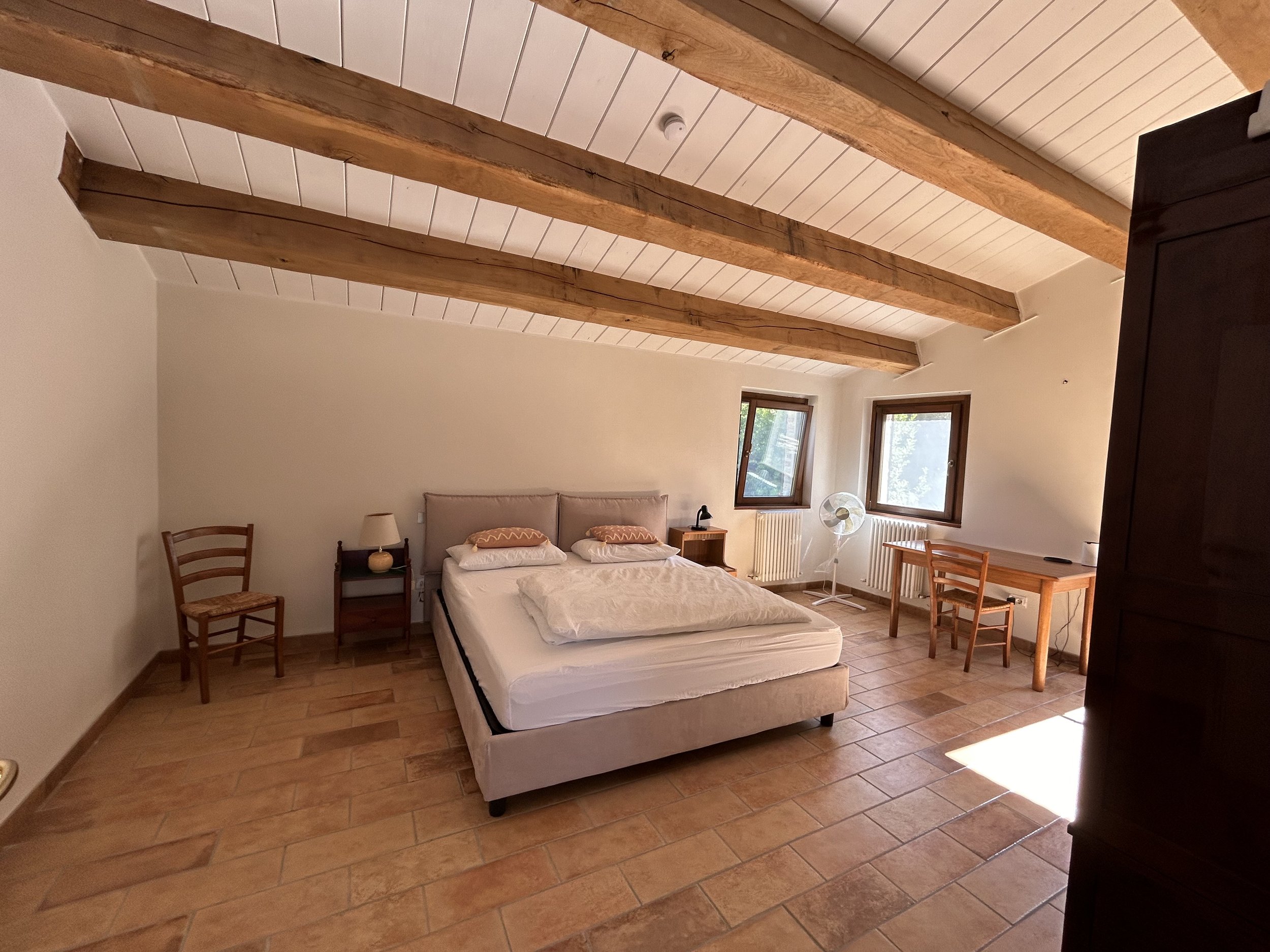 A bedroom with a sloped ceiling featuring exposed wooden beams, a bed with a light-colored headboard and white bedding, a small nightstand with a lamp, a chair, two windows, a fan, a desk with a chair, and tiled flooring.