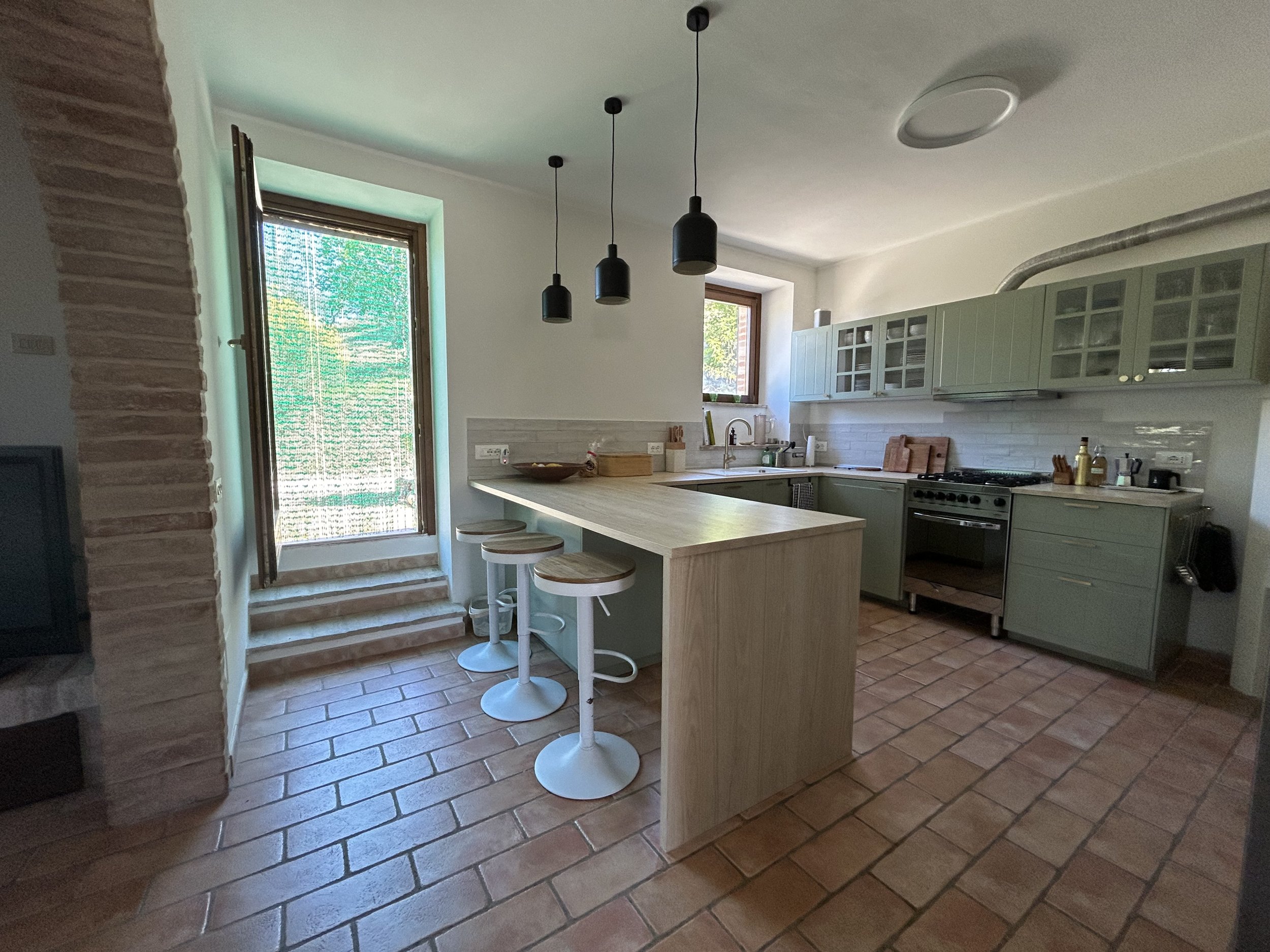 Modern kitchen with a light green cabinetry, a central island with stools, brick flooring, and large windows providing natural light.