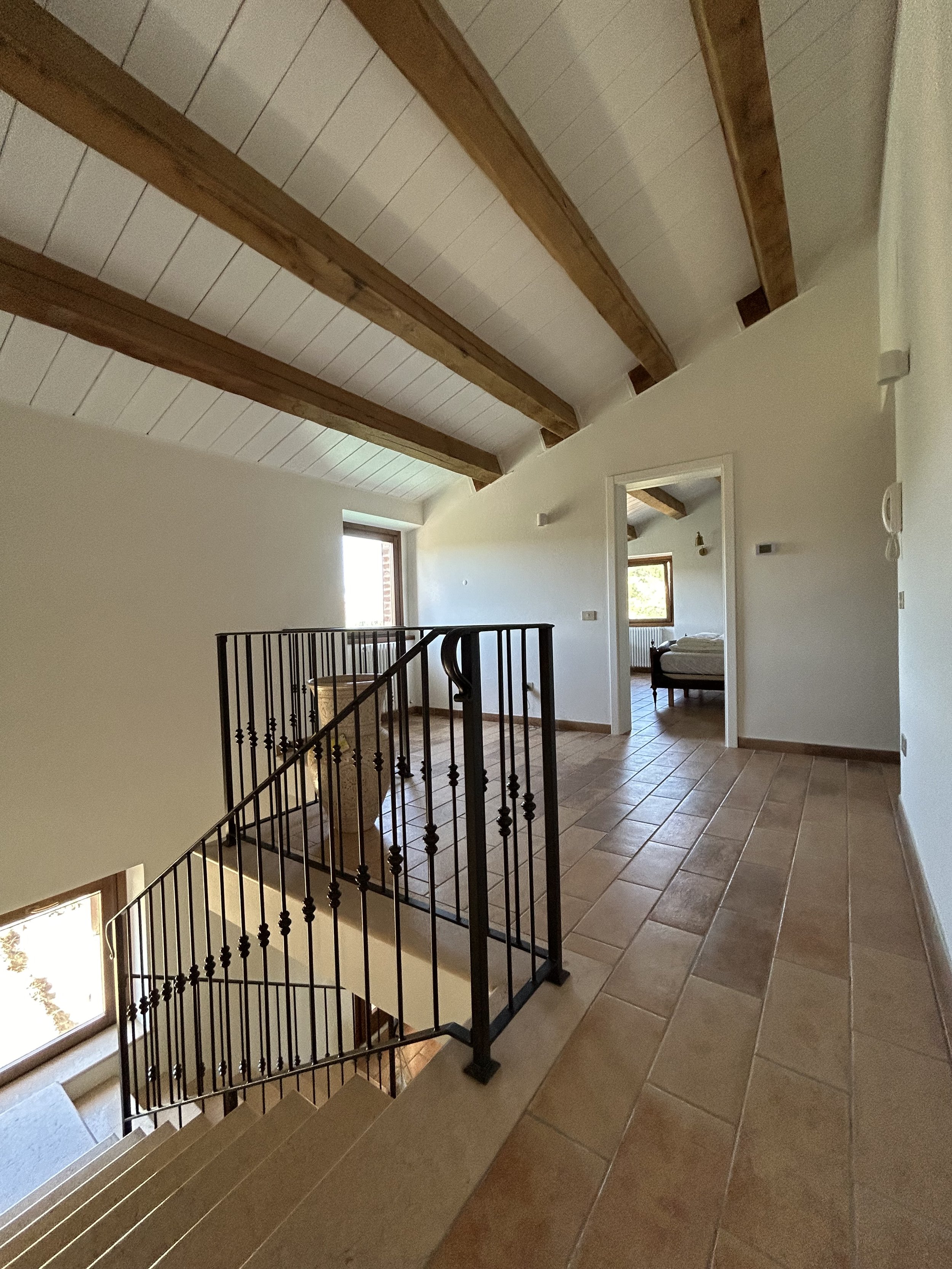 Interior view of a residential staircase landing with tiled flooring, wooden ceiling beams, and a bedroom visible through an open doorway.