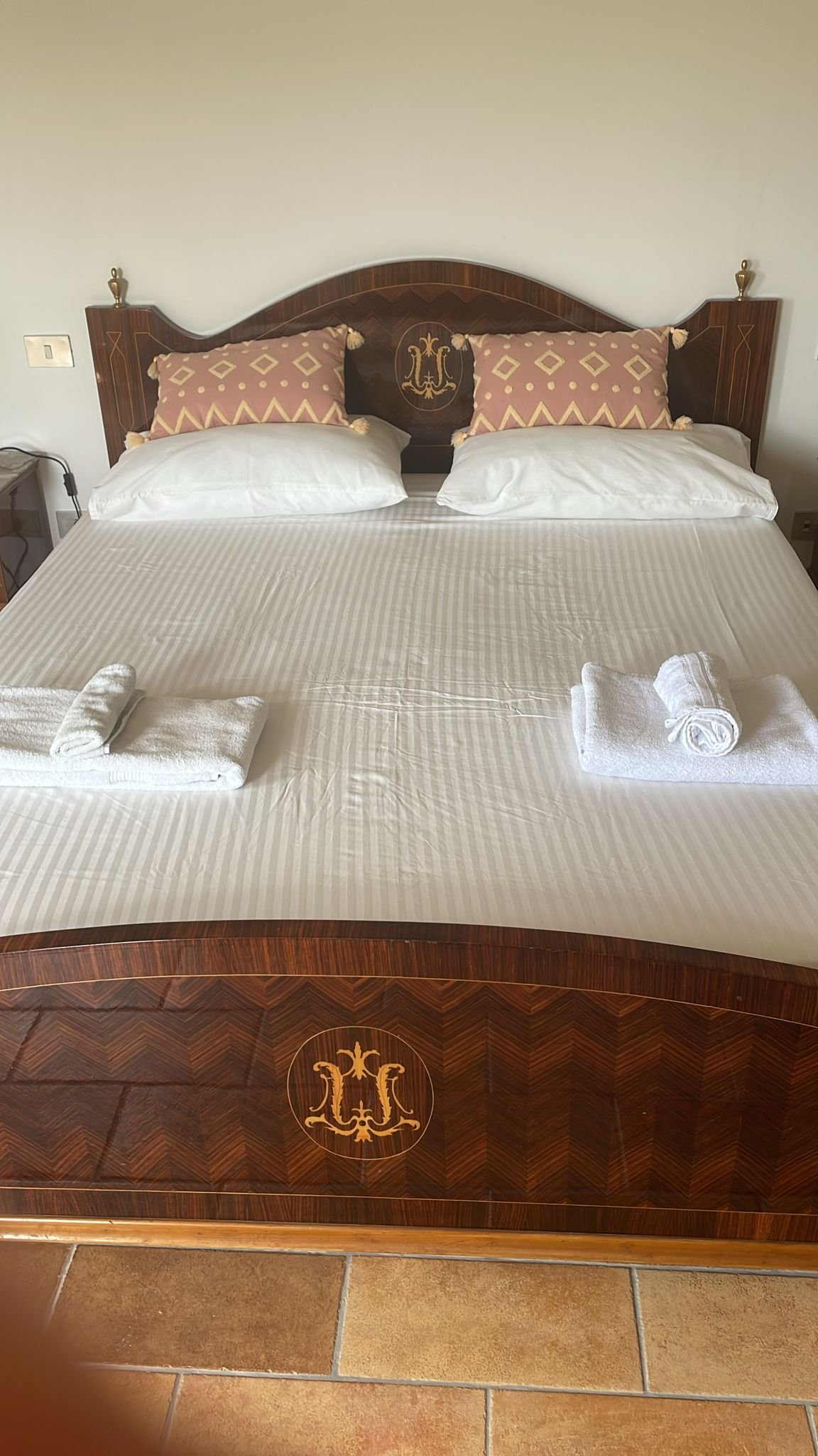 A neatly made hotel bed with white sheets, two neatly rolled towels, and decorative pillows with tassels. The headboard is wooden with ornate inlay and a crest emblem, set against a plain white wall.