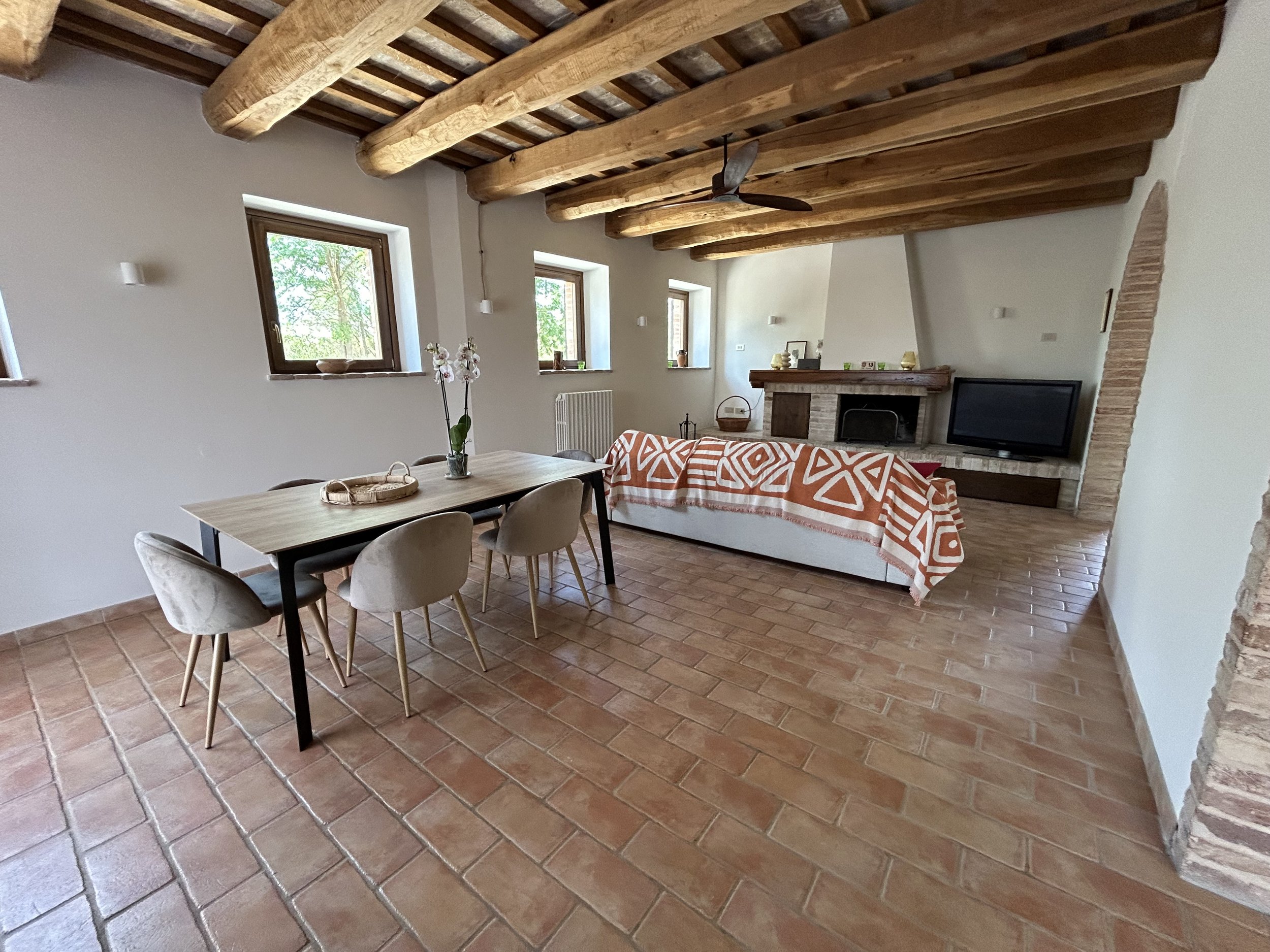Living room with terracotta tile floor, exposed wooden ceiling beams, a dining table with six beige chairs, a white sofa with an orange and white patterned throw, a fireplace, and a flat-screen TV.