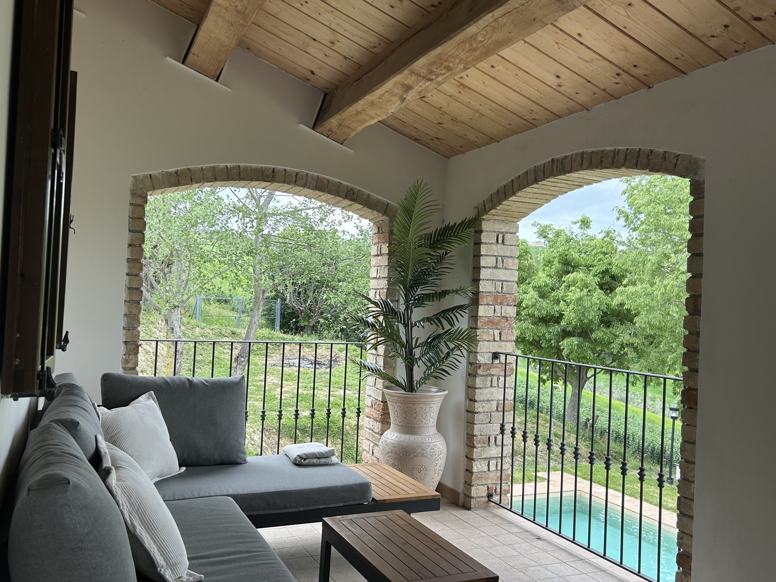 Indoor porch with grey sofa, potted plant, brick arches, wooden ceiling, and a view of trees and a swimming pool outside.