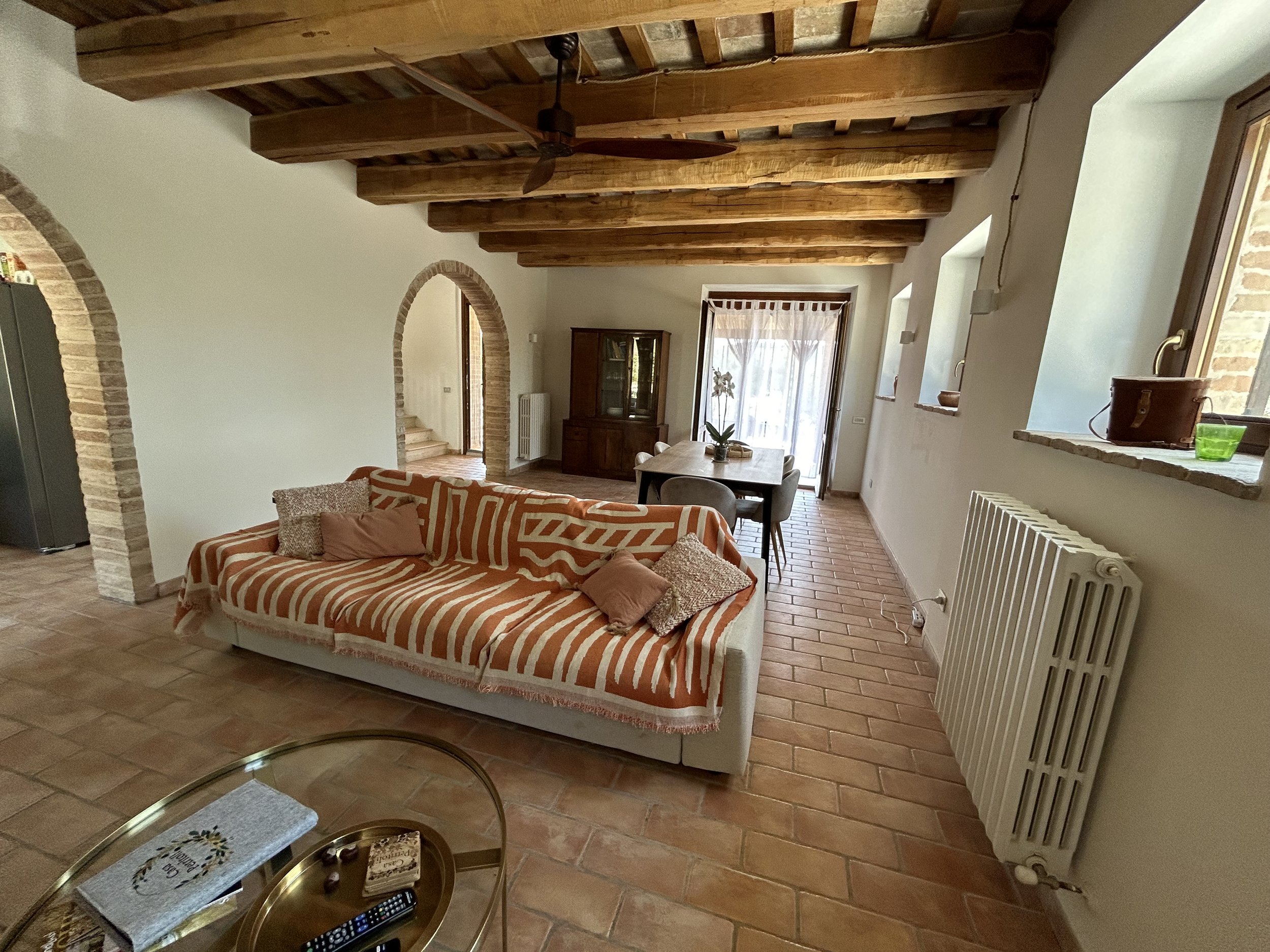 Living room with wooden ceiling beams, a sofa with striped orange and white cover, a glass coffee table, and a dining area with a wooden table and chairs. There are brick arches, windows with ledges, and terracotta tiled flooring.