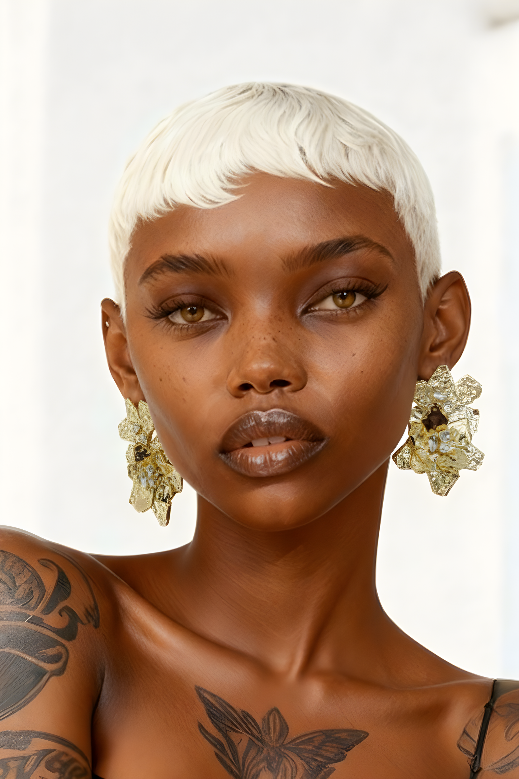 Close-up of a woman with short platinum blonde hair, gold floral earrings, tattoos on her shoulder, and a neutral facial expression.