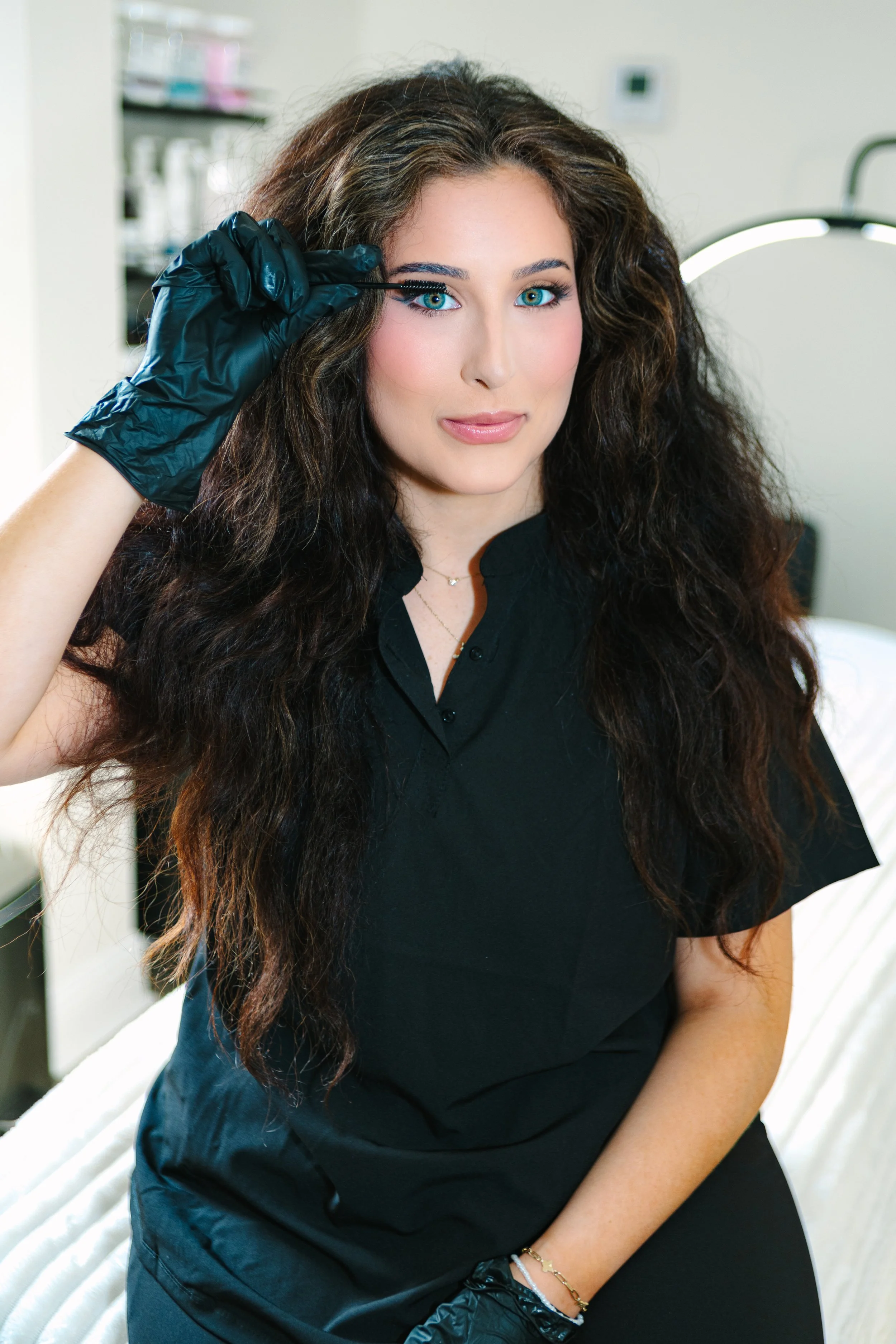 A woman with long dark curly hair, blue eyes, wearing black gloves, and black clothing, applying makeup with a brush in a medical or beauty treatment setting.