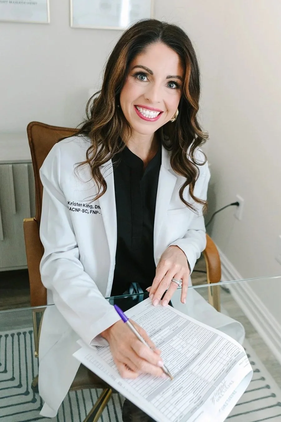 A woman with long wavy brown hair, wearing a white coat and black shirt, smiling while signing a document at a glass table in an office setting.